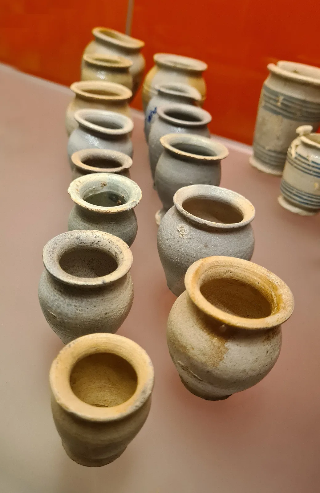 A row of small plain stoneware pill jars in yellow and grey, undecorated, several chipped at the rim and discoloured inside, arranged on a museum shelf