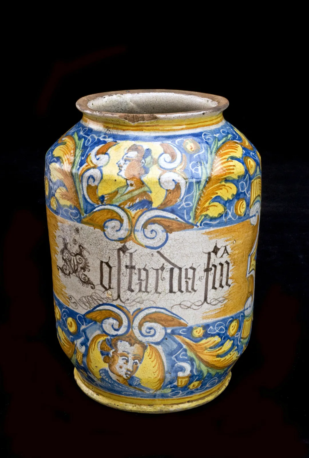 Sicilian tin-glazed drug jar with grotesque masks in yellow and blue and a white band lettered in gothic blackletter, the whole glazed surface covered in a fine irregular network of crazing