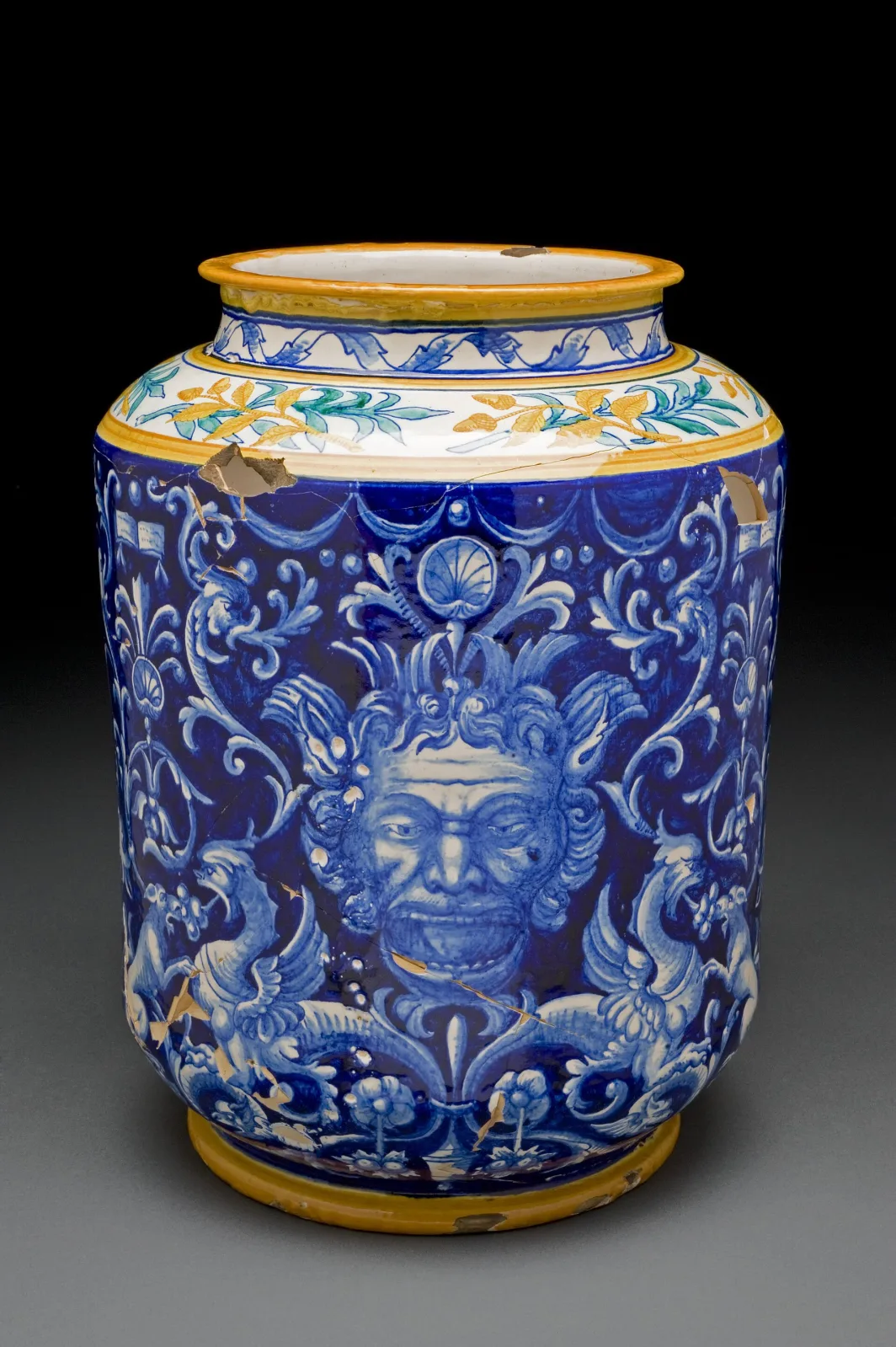 Italian maiolica pharmacy jar with a grotesque bearded mask on a deep blue ground, a foliate band at the shoulder, and a chip at the rim exposing the dark body beneath the glaze