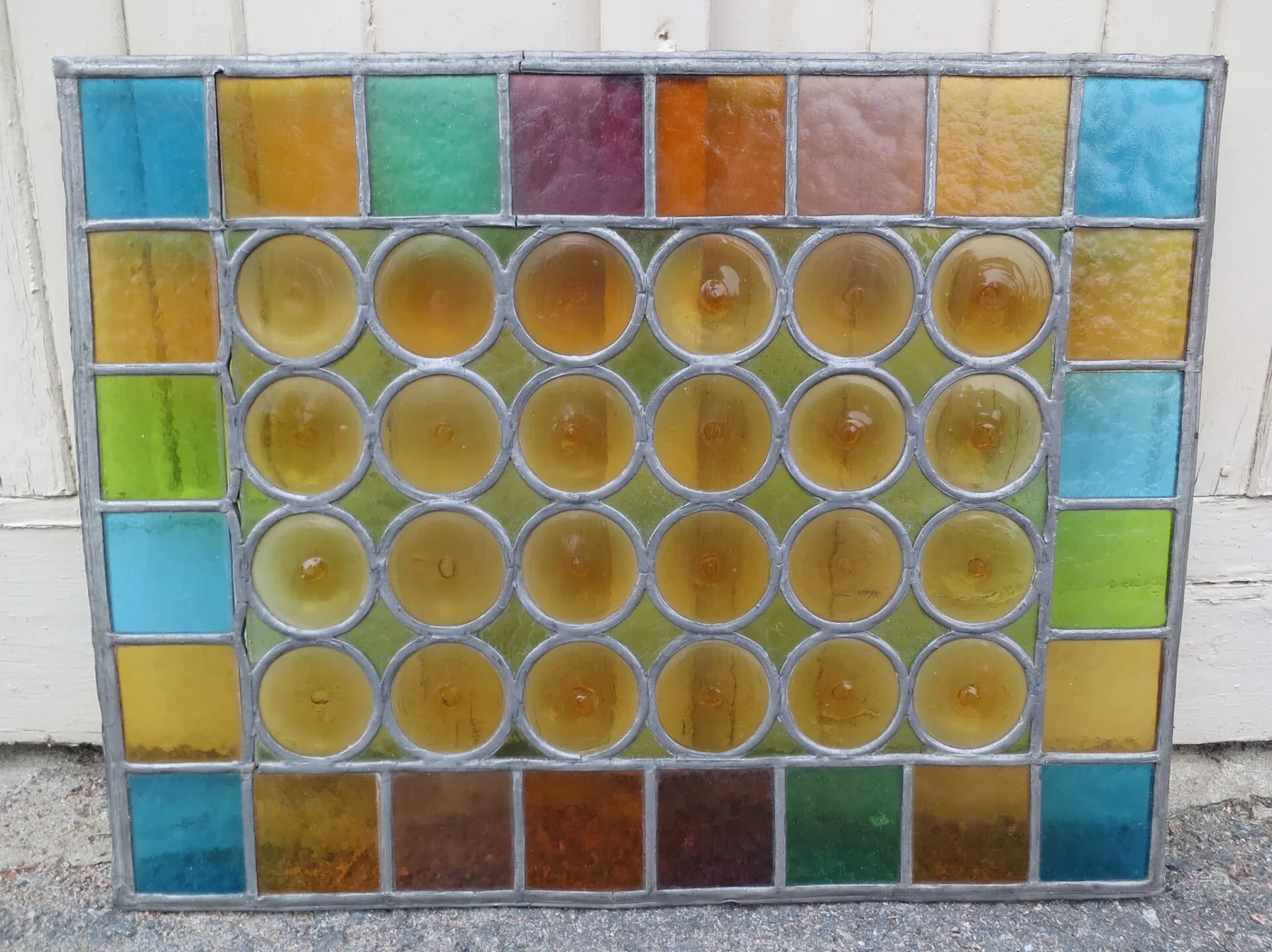 Salvaged leaded glass panel standing on a pavement against a wall, its centre filled with amber bullseye rondels inside a border of coloured squares