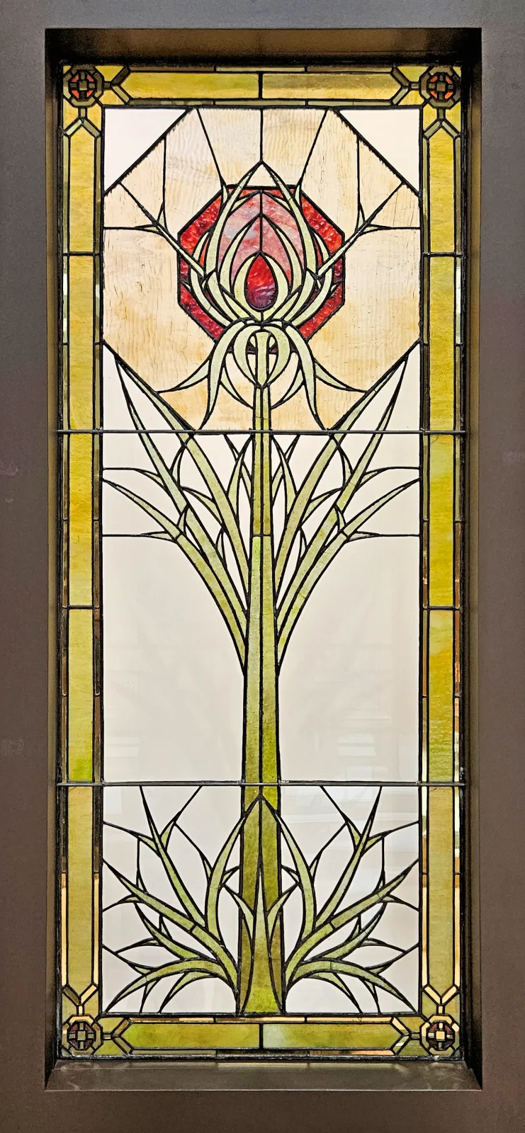 Tall leaded glass window with a stylised thistle rising the full height, in green, cream and red glass within a geometric border