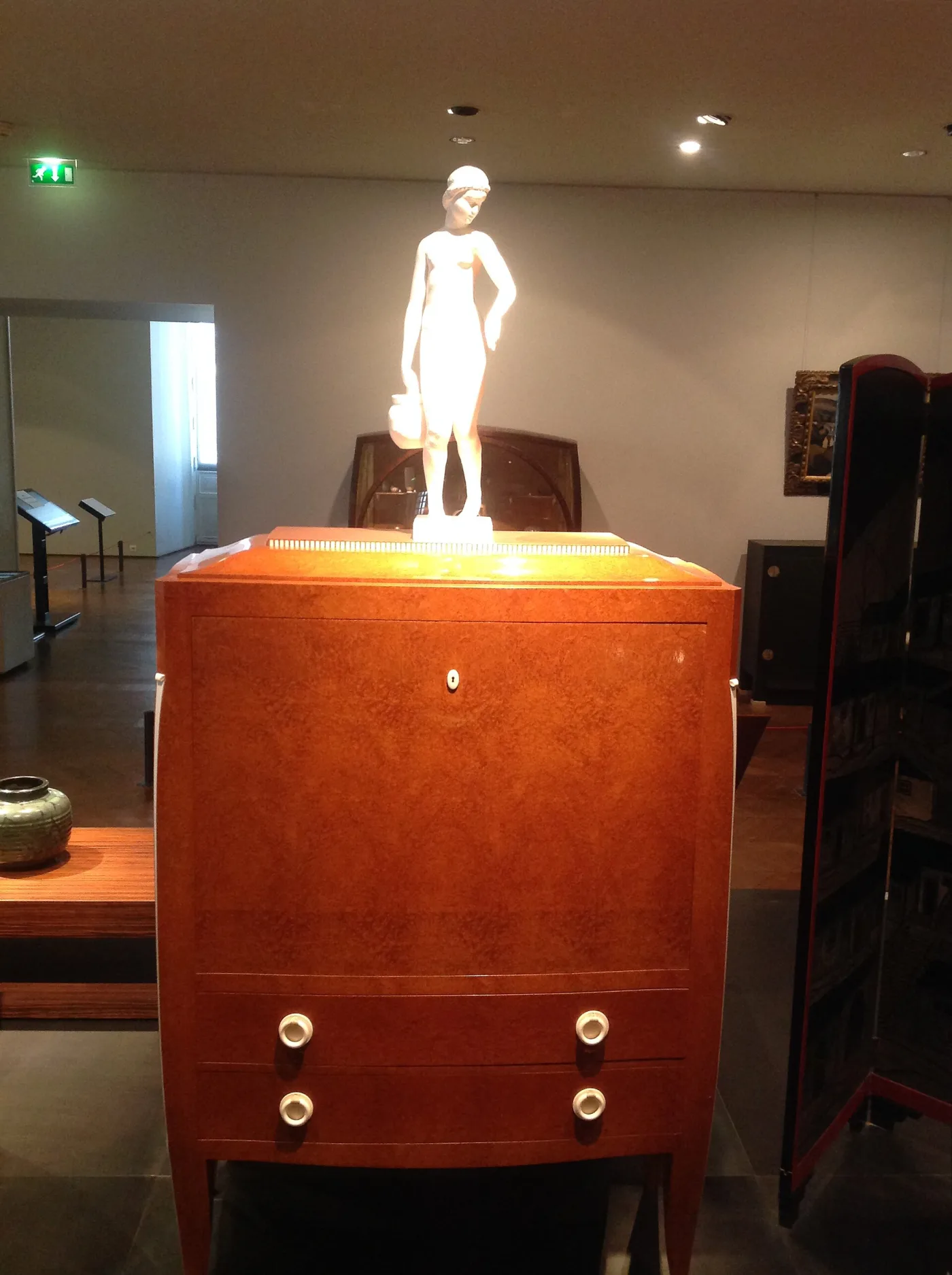 Burl-veneered fall-front secretaire with two drawers and pale circular knobs, a carved figure standing on top