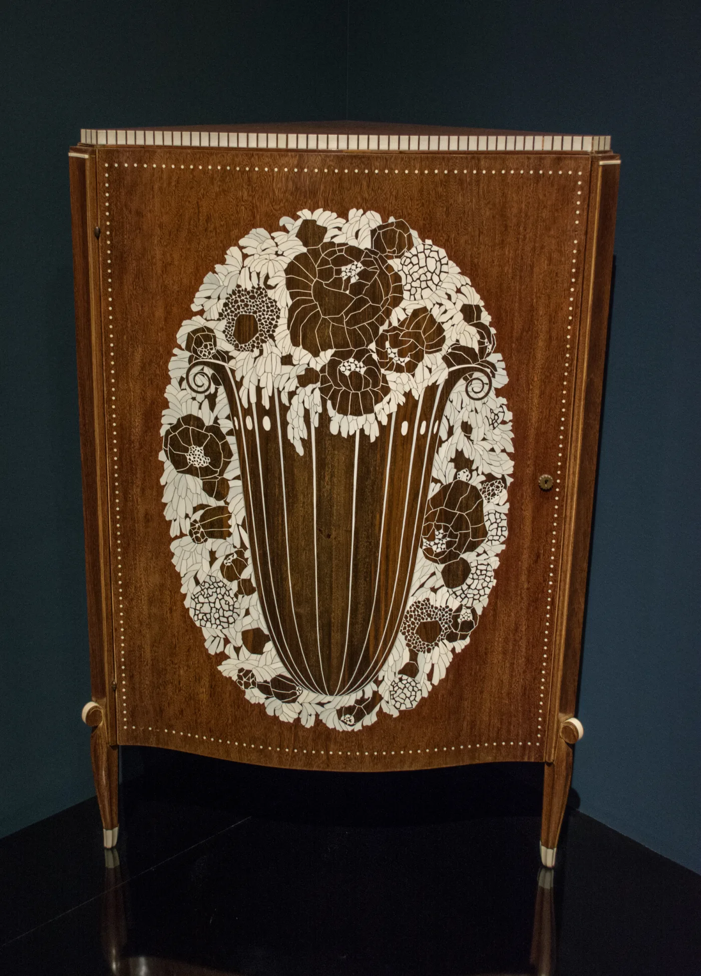 Corner cabinet veneered in dark wood, the door inlaid with an oval ivory basket of flowers, on tapered legs tipped with ivory