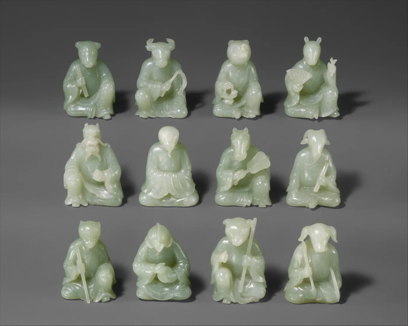 Twelve small pale green jade figures arranged in three rows, each a seated human figure holding or accompanied by one of the animals of the Chinese zodiac