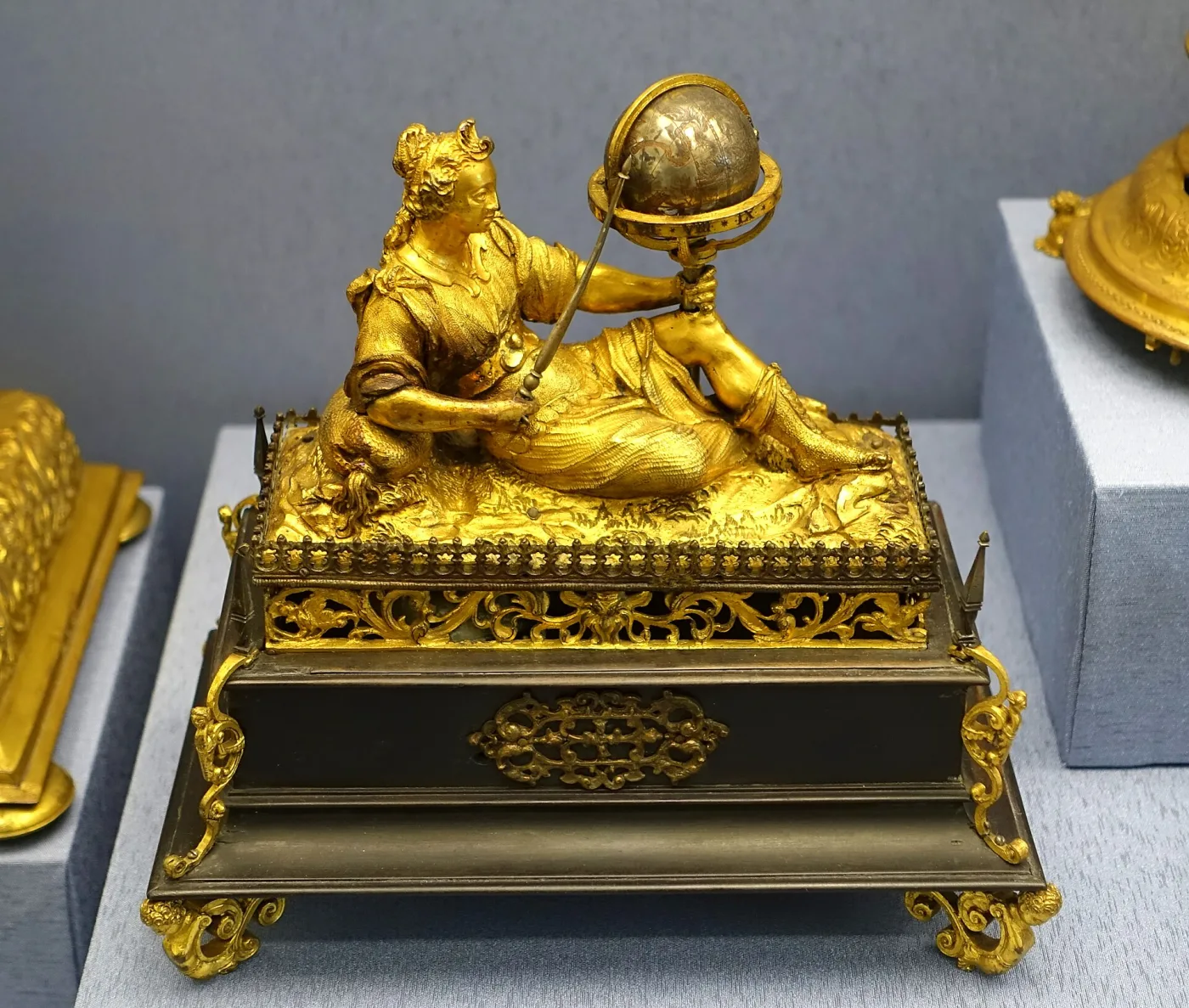 Gilt figure of a reclining woman holding up a small globe, mounted on a dark rectangular clock case with pierced gilt mounts and scrolled feet