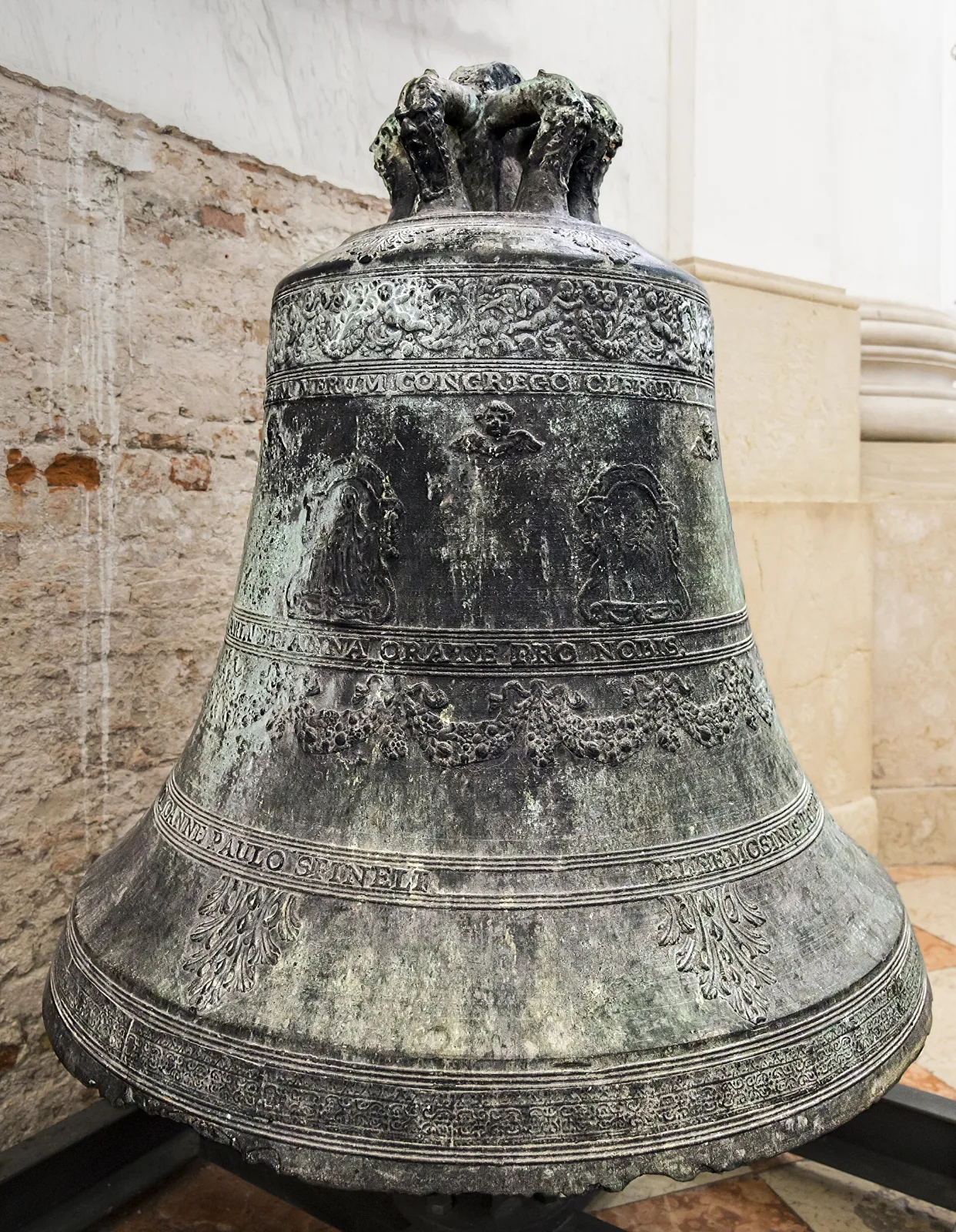 Weathered bronze bell mounted on a timber headstock in a bell chamber, its shoulder carrying bands of relief decoration and lettering, the metal grey-green with age