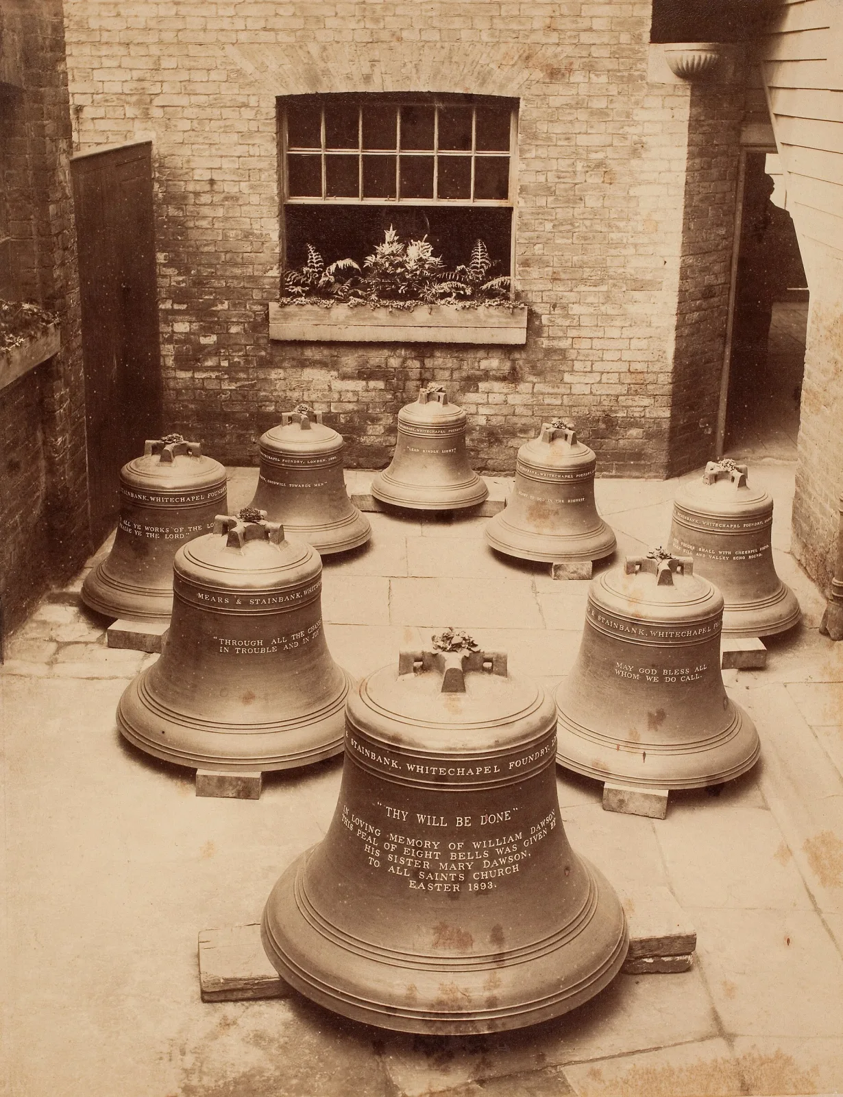 Sepia photograph of a foundry interior with eight newly cast church bells of graduated sizes standing on a brick floor, their shoulders carrying raised inscriptions