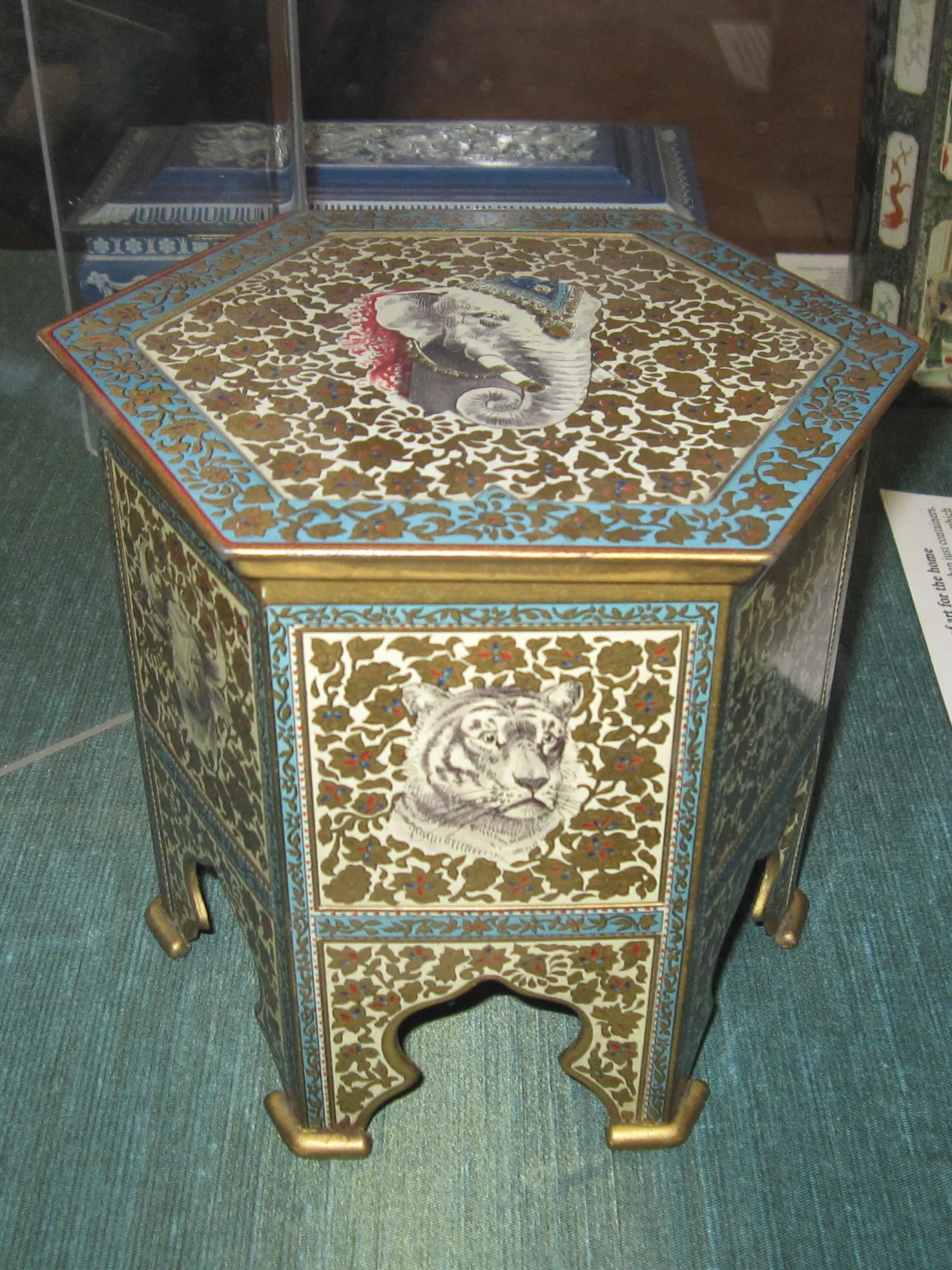 A biscuit tin made in the form of a small Indian occasional table with a decorated top and animal-head ornament