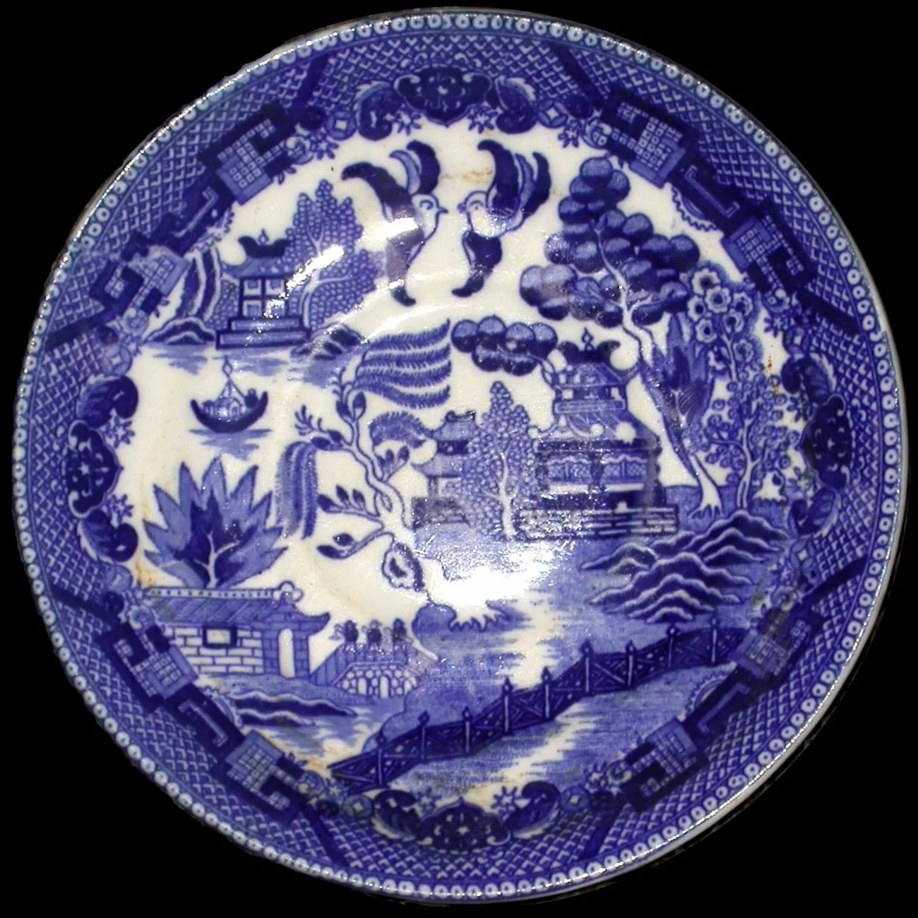 Worn blue and white earthenware plate printed with a willow tree, teahouse, boat, fence and two birds