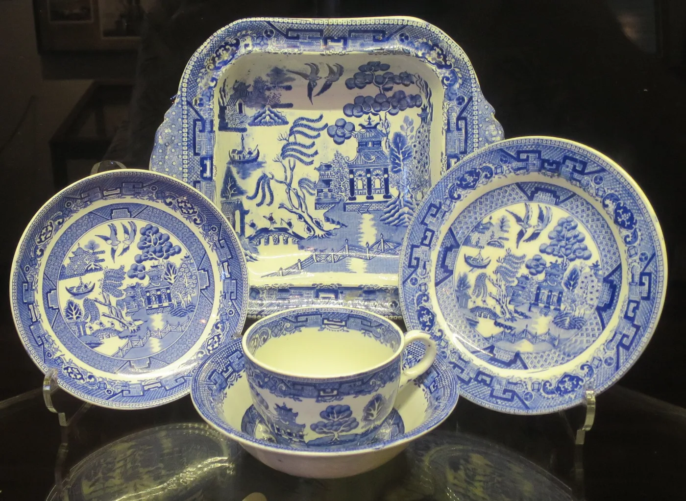 Five pieces of blue and white Willow china grouped in a museum case: a square dish, two plates of different sizes, and a cup and saucer