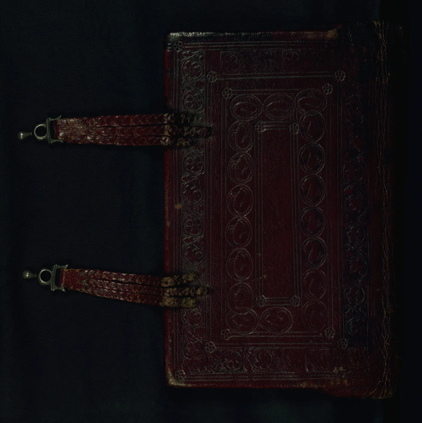 Dark red leather book cover blind-tooled with a panel of foliate ornament and roundels, with two leather straps ending in metal fittings crossing the fore-edge