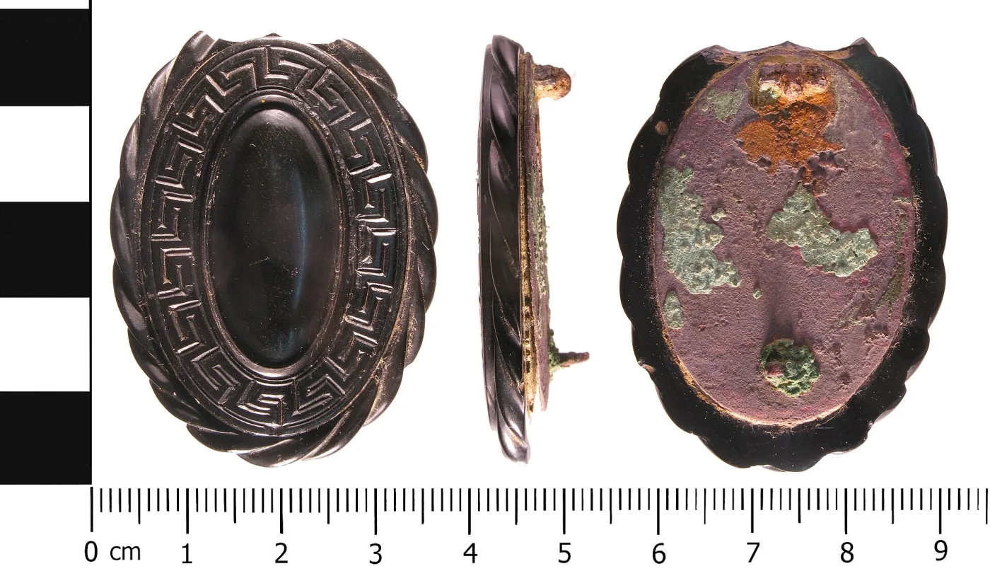 Three views of an oval black mourning brooch beside a centimetre scale: front carved with a Greek key border, edge profile, and back showing a green-corroded metal pin fitting