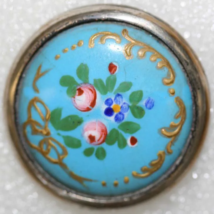 Domed button of opaque turquoise enamel painted with a spray of pink roses and blue flowers and framed by gilt scrollwork, held in a plain metal collet