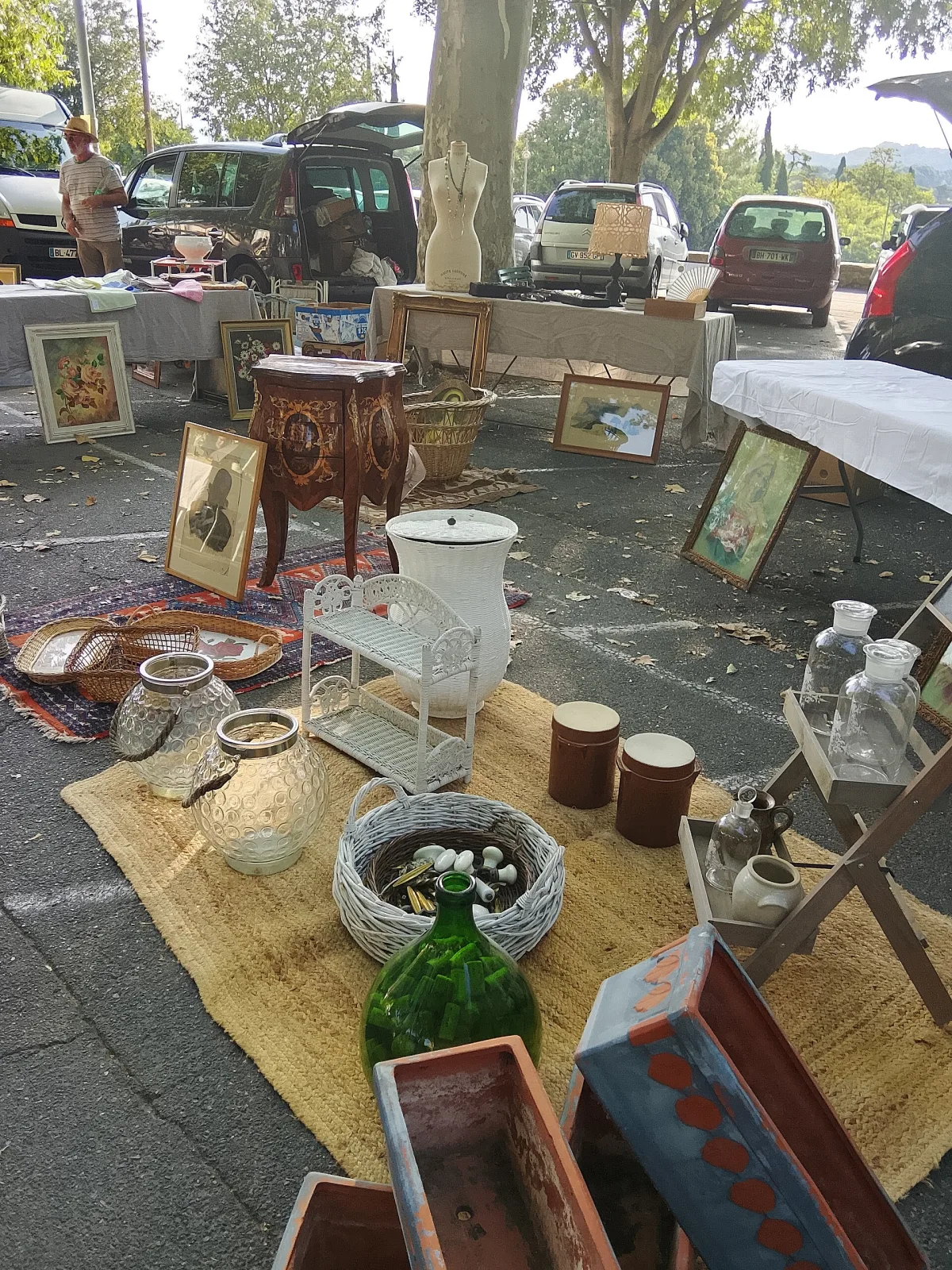 Weekly French brocante laid out on rugs and folding tables in dappled shade under plane trees, with copper pans, glass demijohns and small furniture on the ground