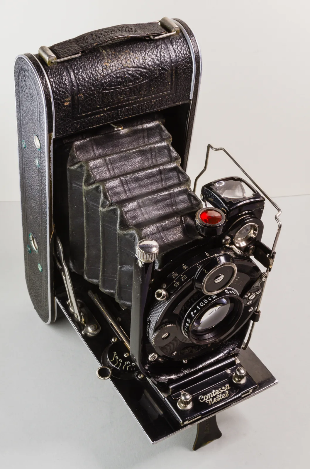 Folding plate camera standing open on its drop bed, the leather-covered body behind a black concertina bellows carrying a round lens in a shutter housing, with a small red-topped viewfinder at the top