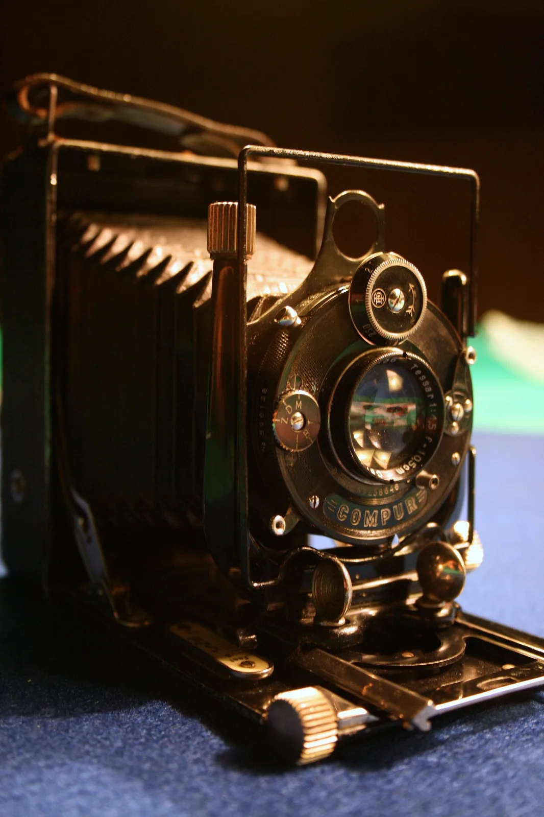 Folding camera opened on its drop bed under warm lamplight, showing a tapered black bellows, a brass-rimmed lens and shutter, and nickel struts holding the front standard