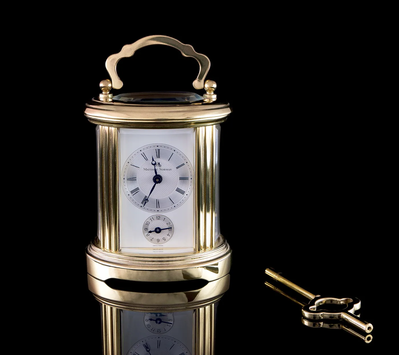 Modern gilt oval carriage clock signed Matthew Norman on a silvered dial, with a subsidiary alarm dial and a double-ended brass winding key beside it