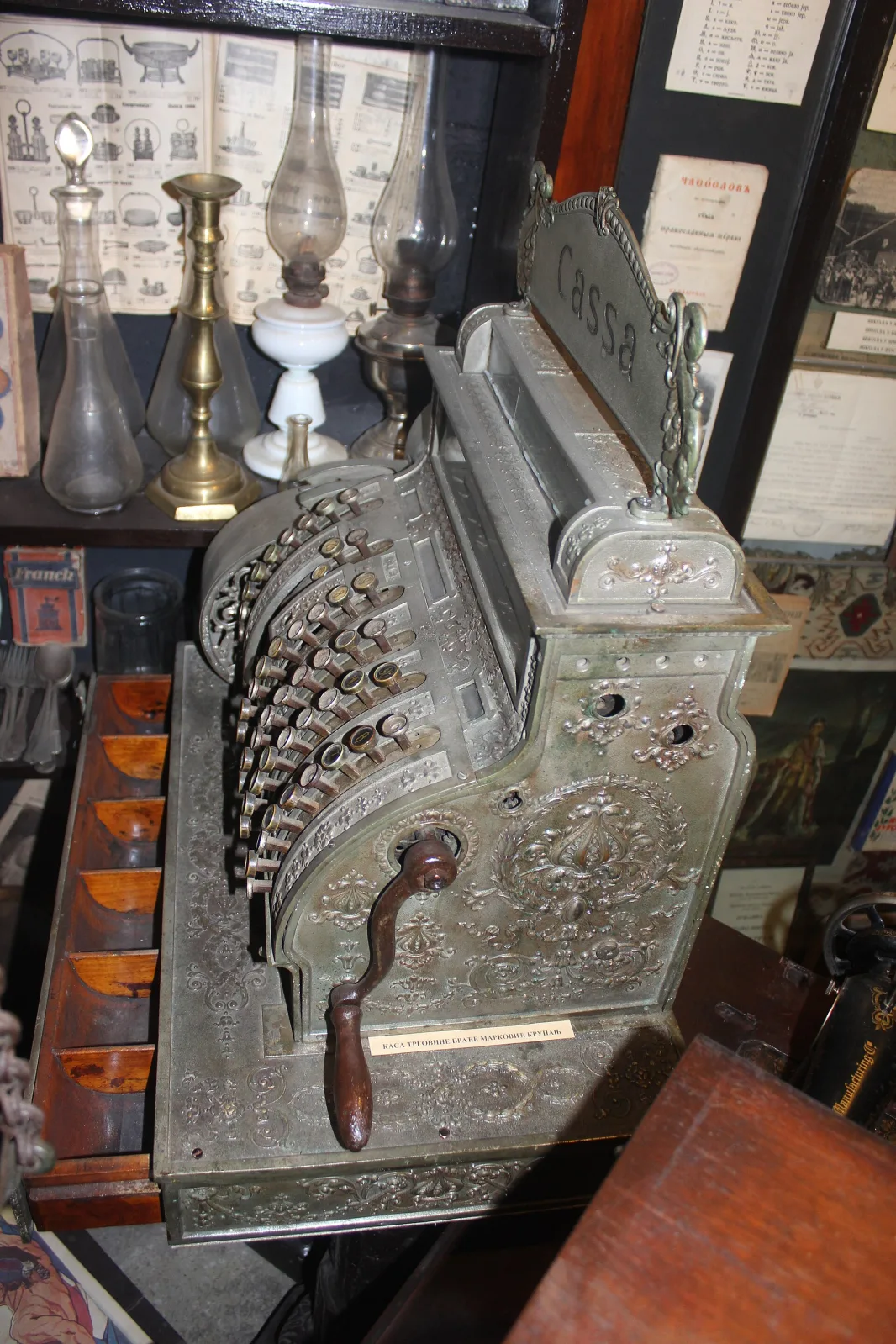 Nickel-plated cash register with a bright reflective case standing among glassware and lamps in a crowded shop