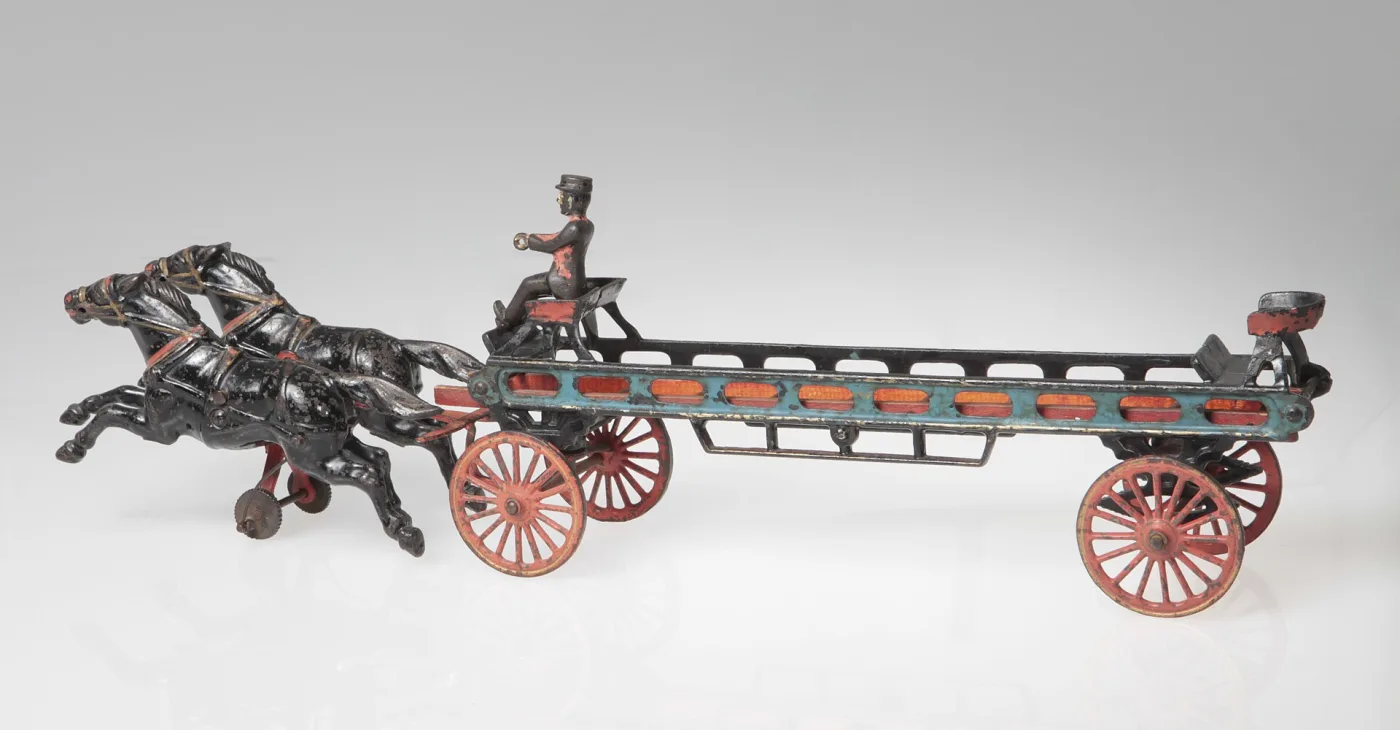 Cast iron toy hook and ladder fire wagon drawn by two white horses, with red spoked wheels, two seated riders and a long ladder carried along the frame