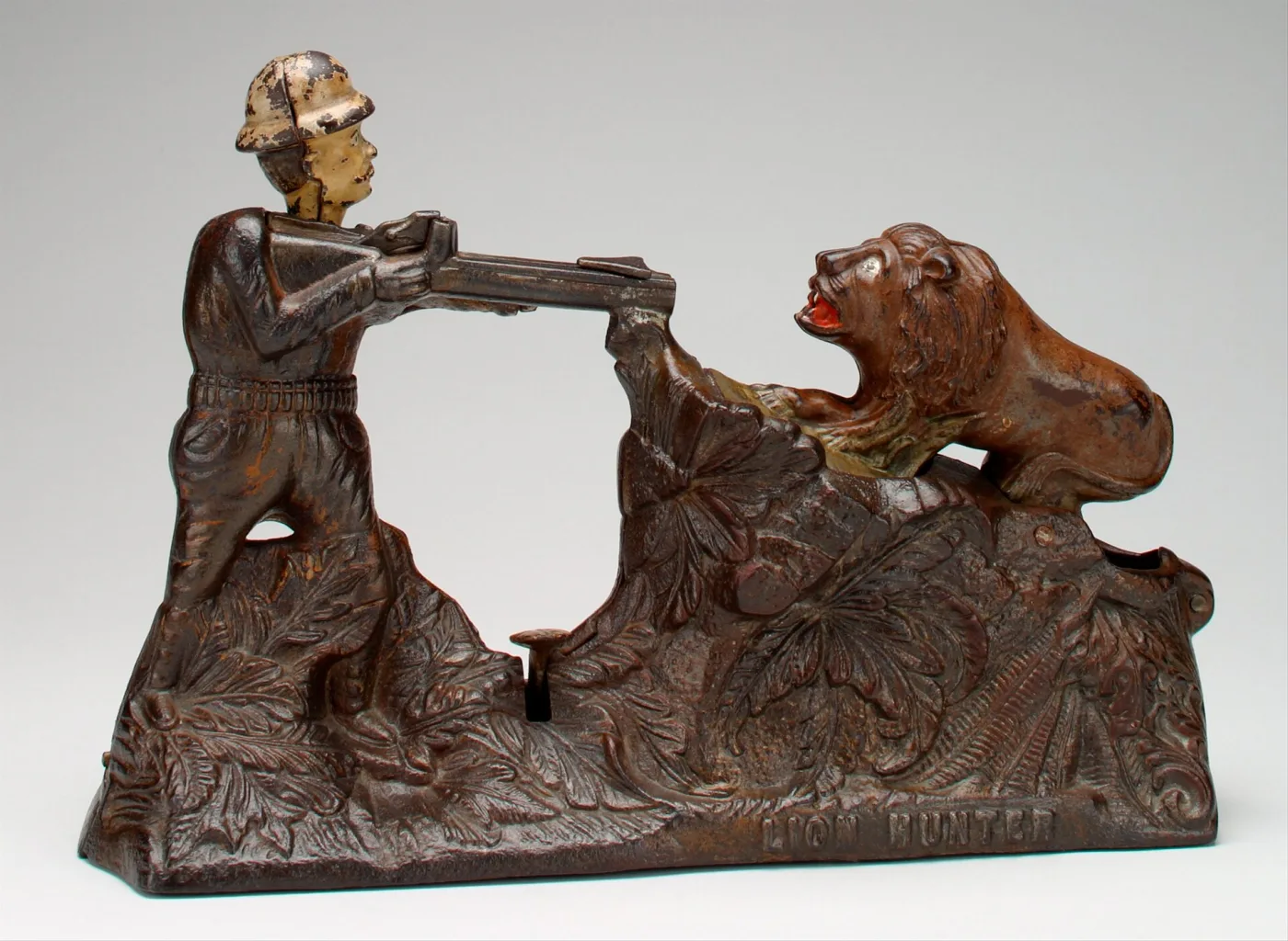 Painted cast iron mechanical bank showing a hunter in a pith helmet kneeling with a rifle aimed at a lion crouched among foliage, on a long rectangular base