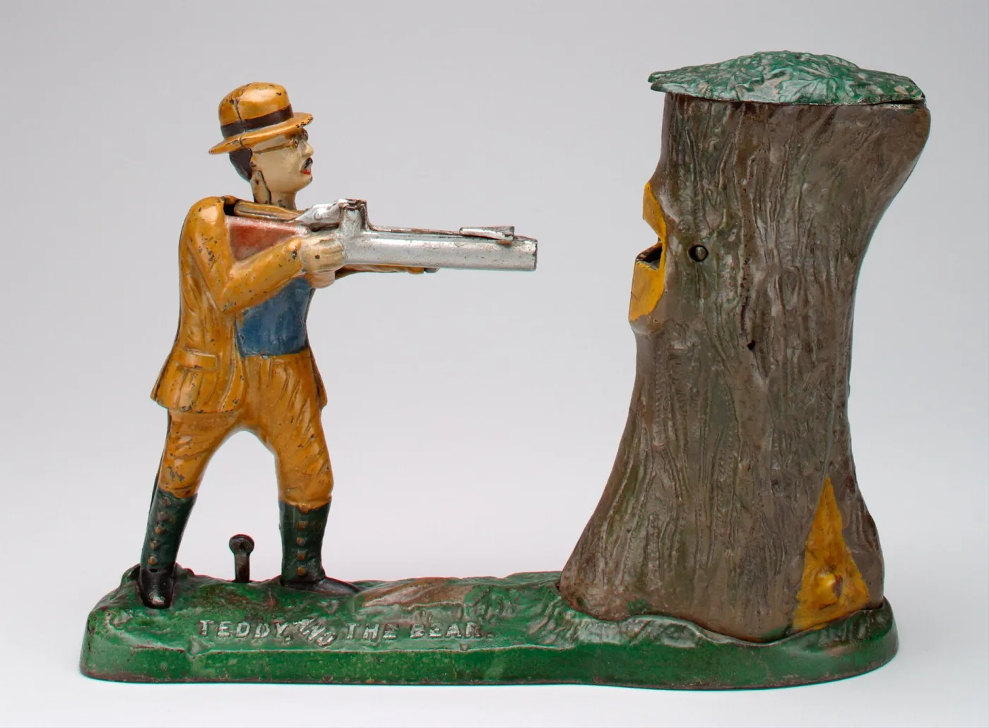 Painted cast iron mechanical bank showing a man in a yellow suit aiming a rifle at a tree stump, with a bear's head emerging from the top of the stump, on a green base