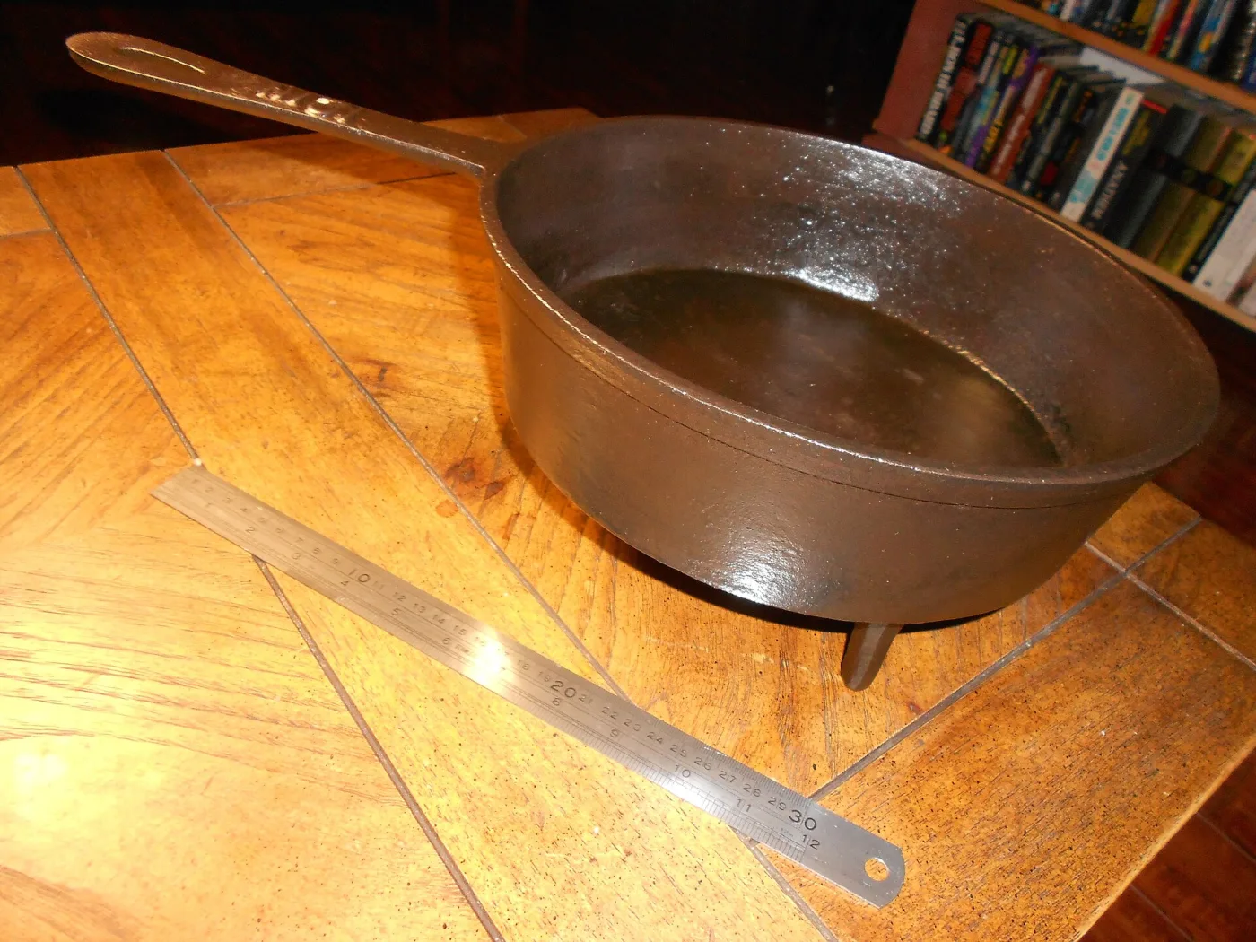 Shallow round cast iron pan on three legs with a long straight handle, standing on a table beside a twelve-inch ruler