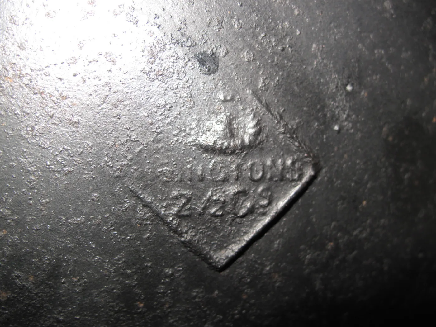 Close view of the underside of a cast iron pot showing a raised diamond-shaped cartouche containing worn lettering and numerals, the surface pitted and encrusted