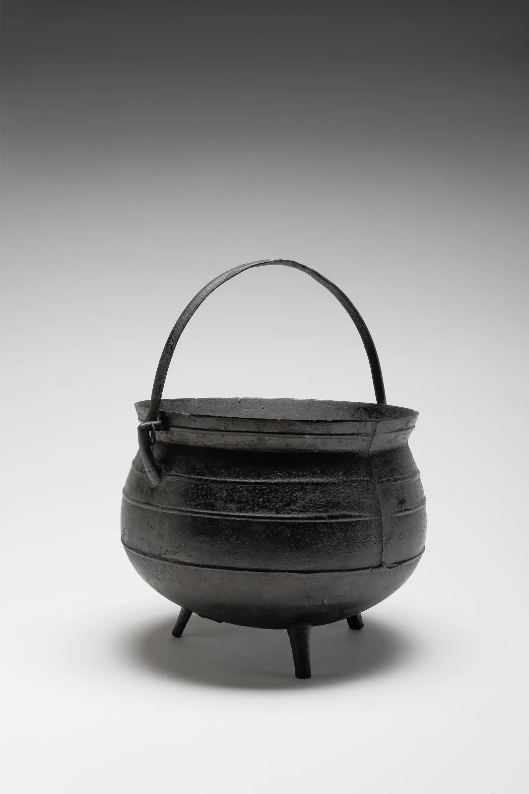 Spherical three-legged cast iron cooking pot with a horizontal band around its body and a swinging bail handle, photographed against a plain graduated background
