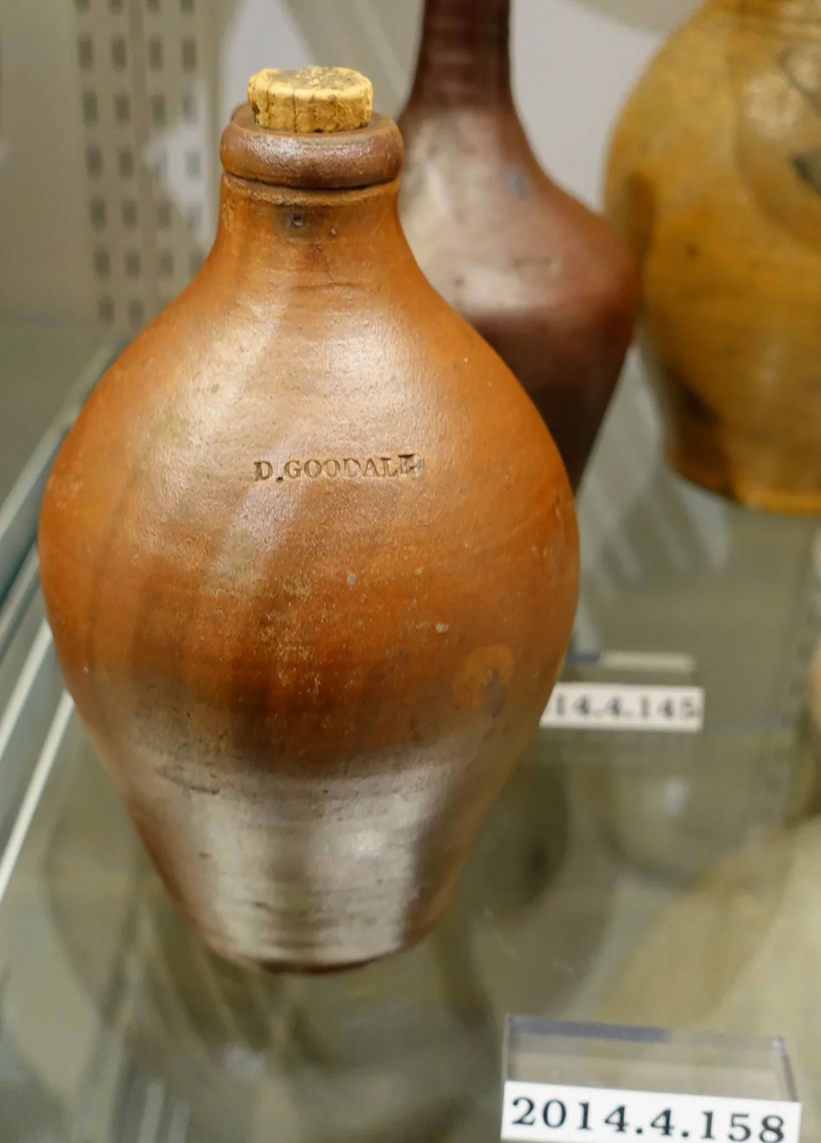 Salt-glazed stoneware jug in a museum case with the name D. Goodale impressed in a line across its shoulder