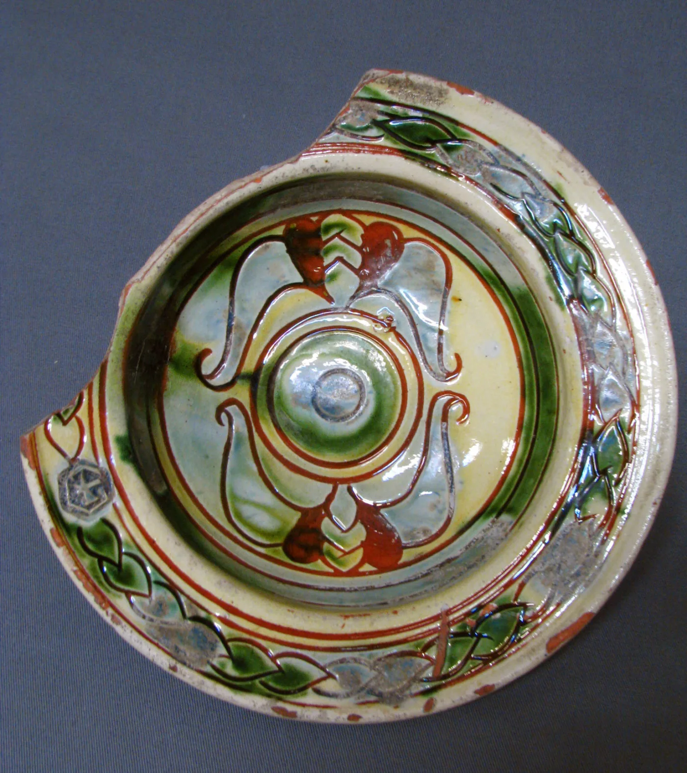Tin-glazed dish seen from above, painted with concentric bands of red, green and blue ornament around a raised central boss, with a wedge broken from the rim