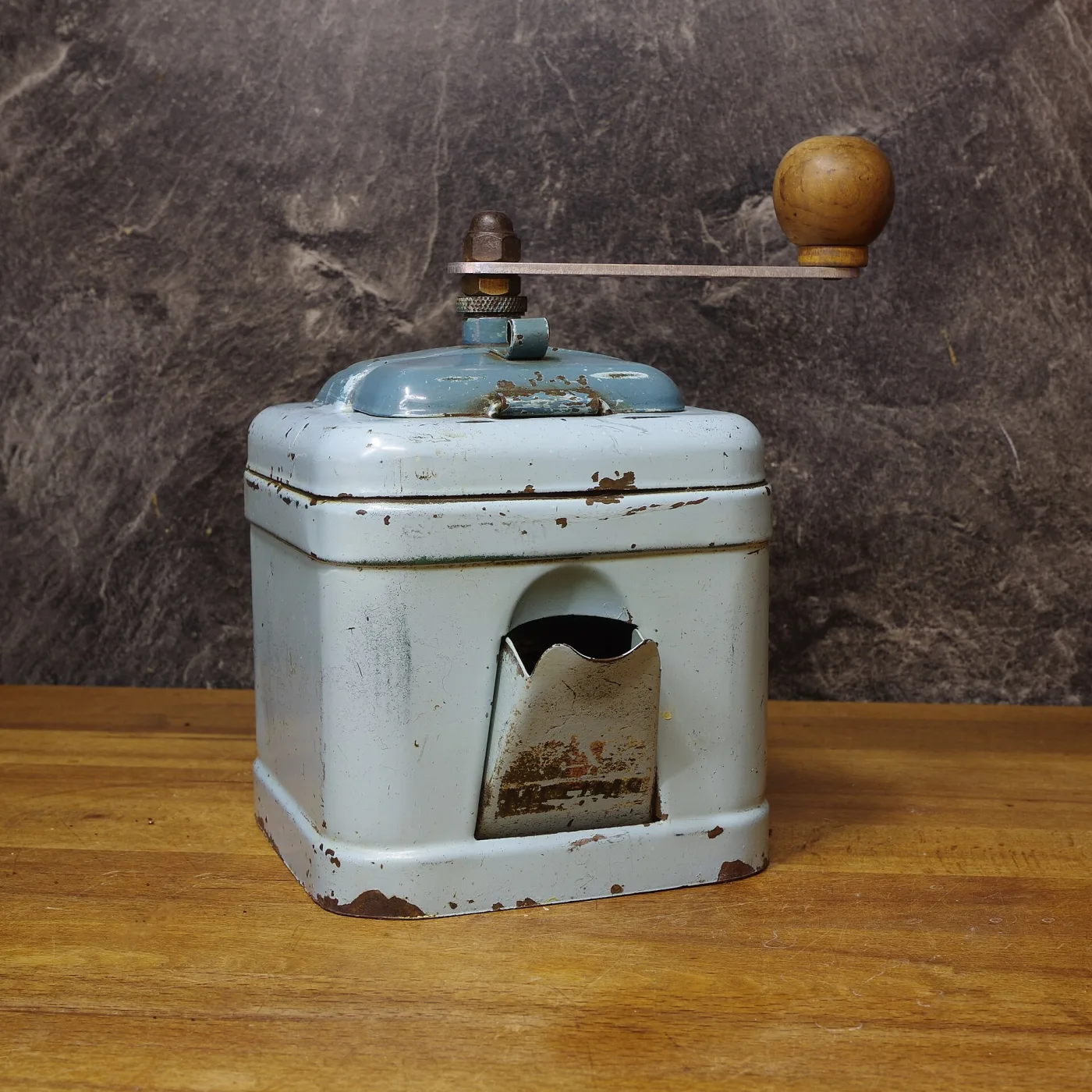 Small tin-bodied coffee mill with a rounded cream body, an open chute at the front and a long crank arm, standing on a dark surface