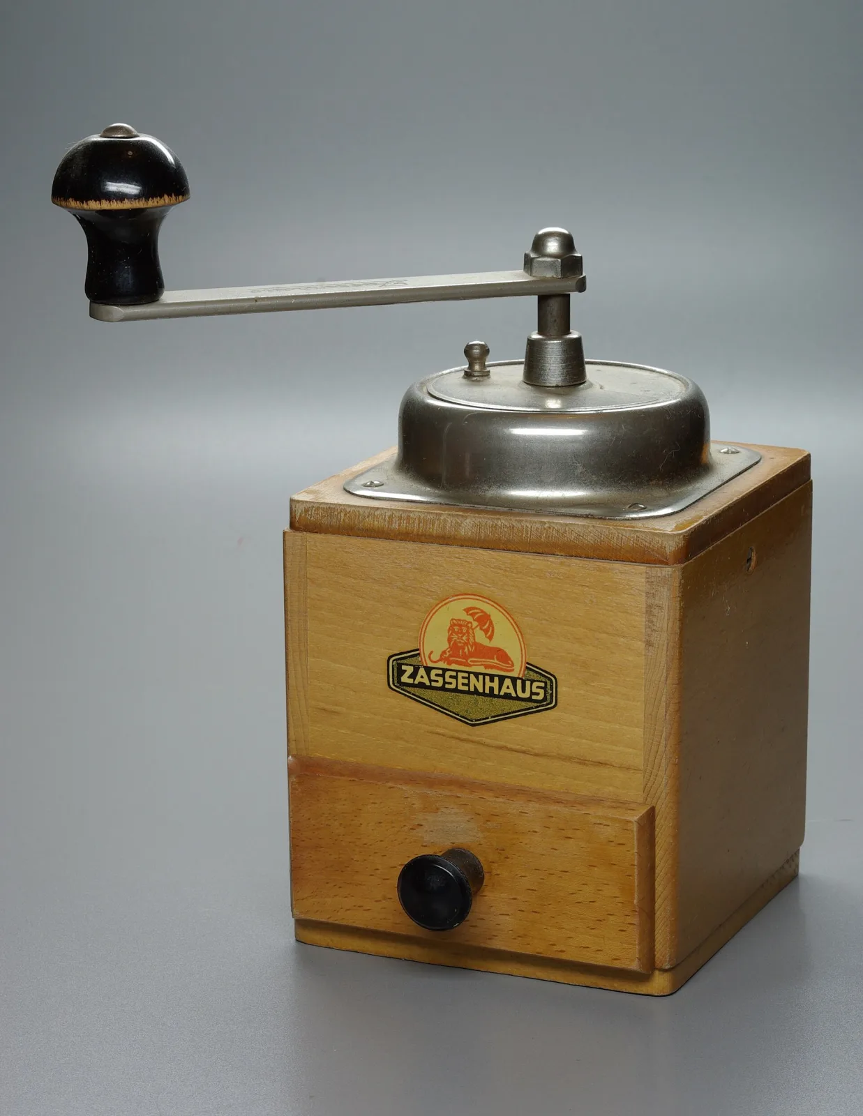 Light wooden box coffee mill with a chromed domed lid and a straight steel crank with a black knob, photographed against a plain grey ground