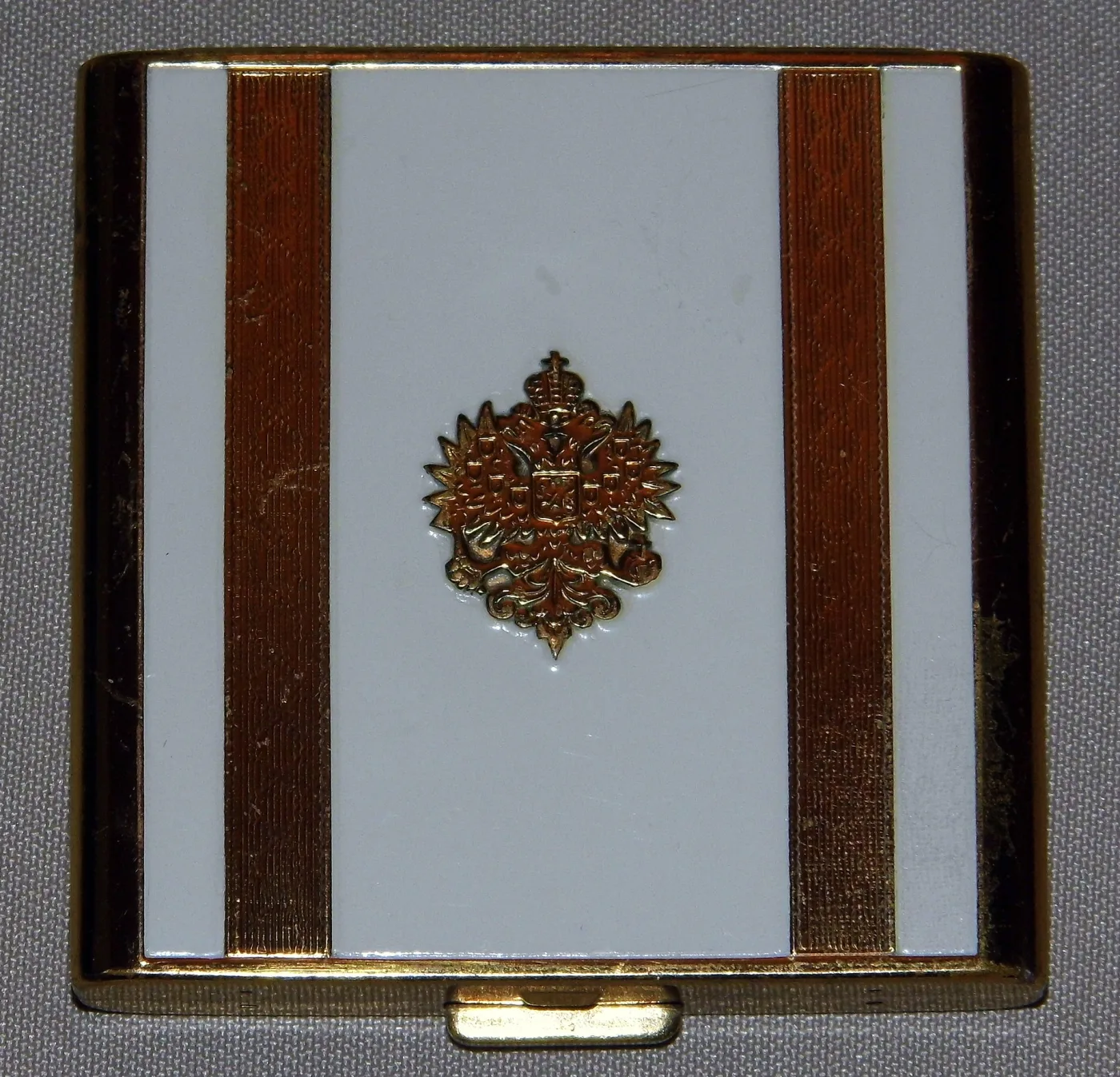Square compact with a pale cream lid divided by two vertical dark brown bands, a small gilt filigree ornament at the centre, in a gilt frame with a push-button catch at the front edge