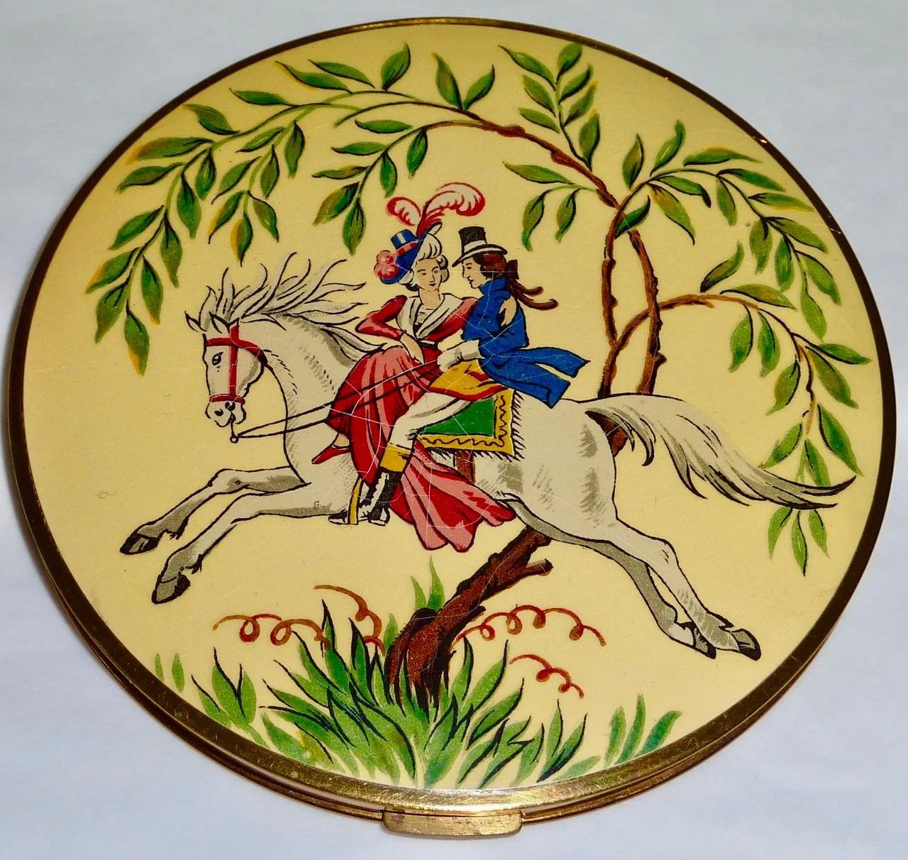 Round compact lid painted with a Regency couple in red and blue riding a white horse beneath a willow, in full colour on a cream ground
