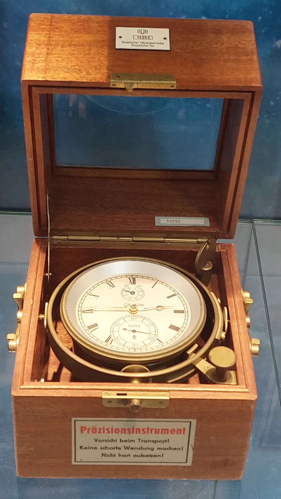 Marine chronometer in a three-tier mahogany box with brass corner plates and a sunk carrying handle, the outer lid raised and the glazed inner lid open above the gimbal-mounted dial, with printed warning labels inside