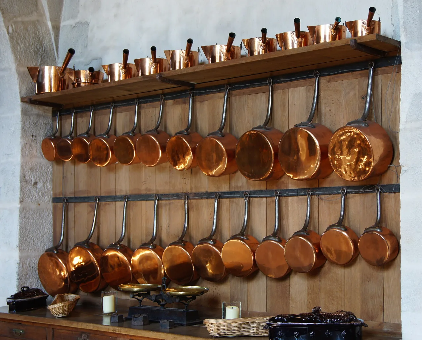 Two long rails of copper saucepans hanging by their iron handles against a boarded wall, graduated from small to large, with a shelf of copper measures and pots above