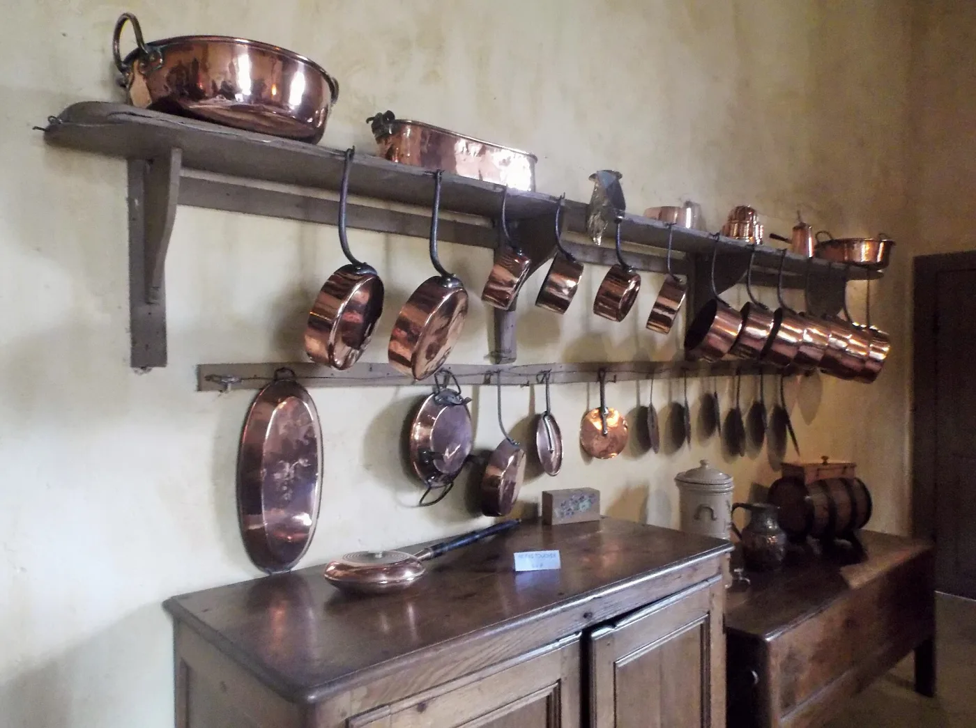 Copper pans of graduated sizes hanging beneath a wooden shelf in an old French kitchen, with a large copper cauldron, a lidded pot and other vessels ranged along the shelf and a wooden dresser below