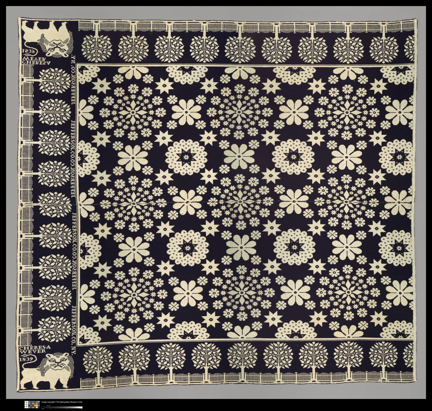 Large coverlet in dark indigo and undyed white, its field filled with rows of flower medallions and eight-pointed stars, with borders of trees behind a fence along the top and bottom, a column of woven lettering down the left side, and a woven lion above a lettered block at each left corner