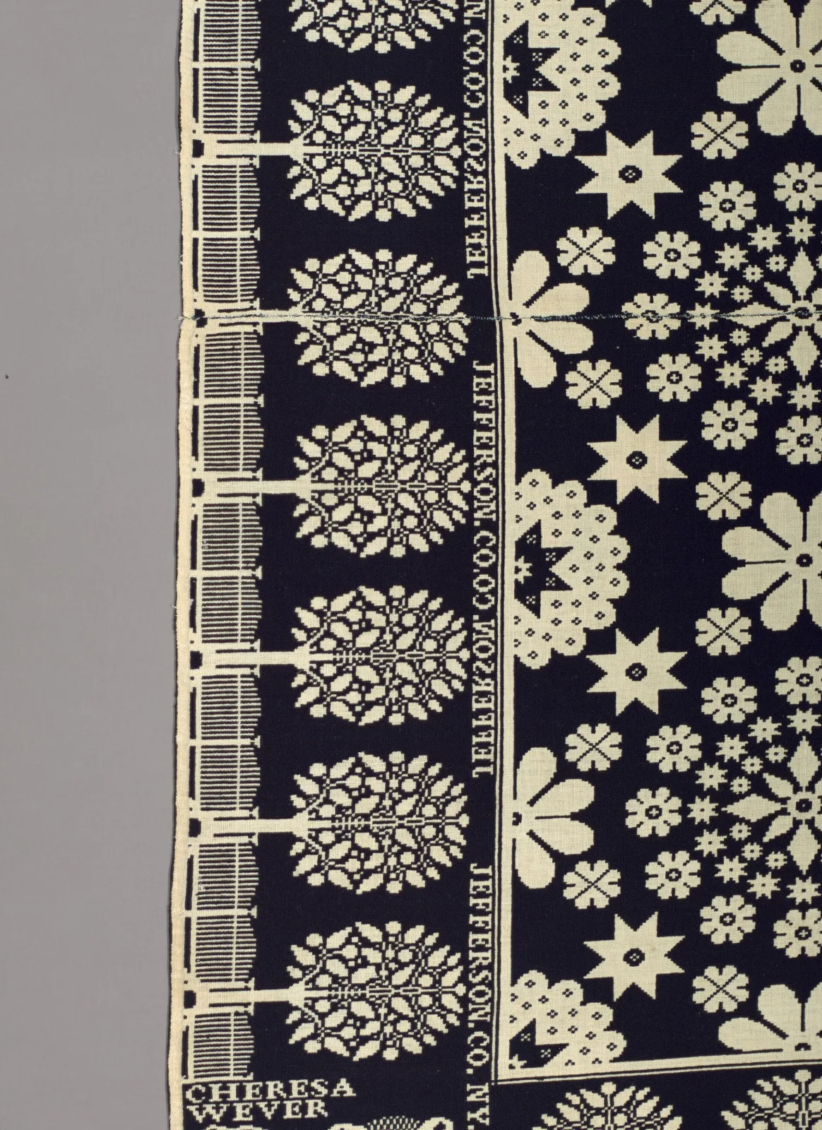 Detail of a dark indigo and white coverlet where the words JEFFERSON CO. N.Y. are woven repeatedly down a narrow vertical border beside a column of flower medallions, with a name legible at the lower edge