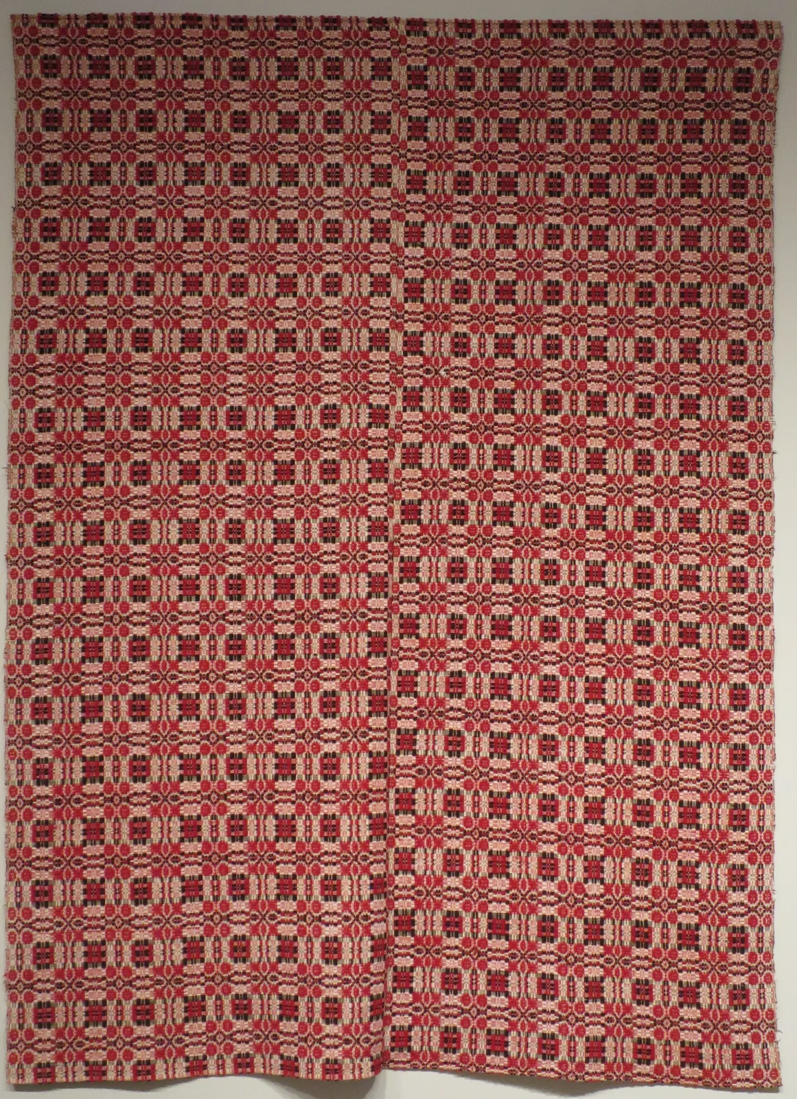 Coverlet woven in dark red and undyed white in a strict grid of small squares and crosses, entirely rectilinear with no curved lines anywhere, and a vertical seam running down the centre where two woven panels are joined