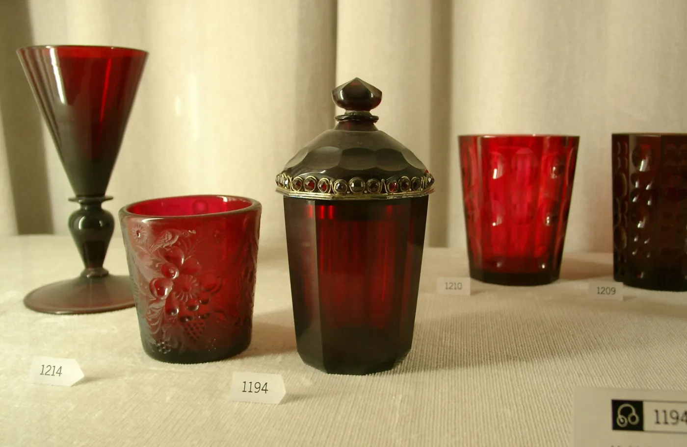 Five deep red glass vessels of different shapes in a museum case, ranging from a bright red tumbler to a faceted covered beaker so dark it reads almost black, one with a jewelled gold mount
