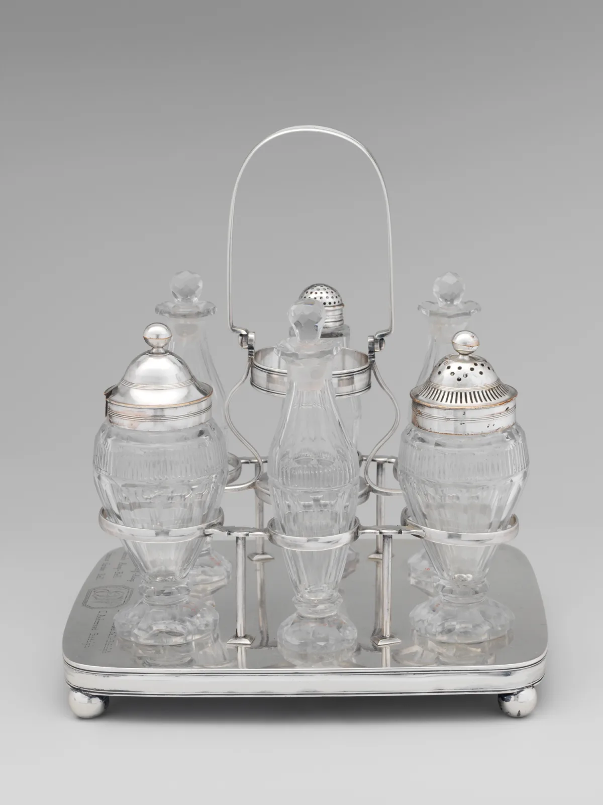Silver cruet frame on a rectangular ball-footed base with a tall arched carrying handle, holding six cut-glass vessels: three tall bottles with faceted stoppers, two ovoid jars with pierced silver caster tops and one with a plain domed silver cover