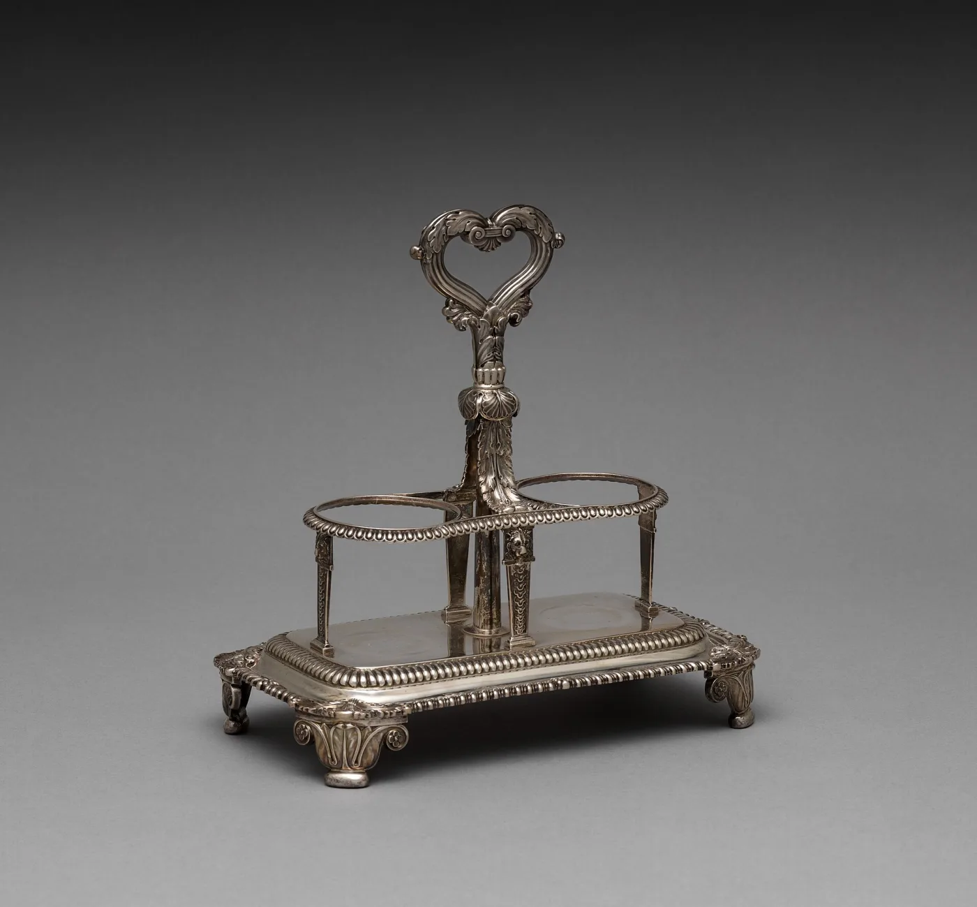 Empty silver two-ring cruet frame with gadrooned rims, a heart-shaped foliate handle on a central column, standing on a rectangular gadrooned base with four leaf-capped feet