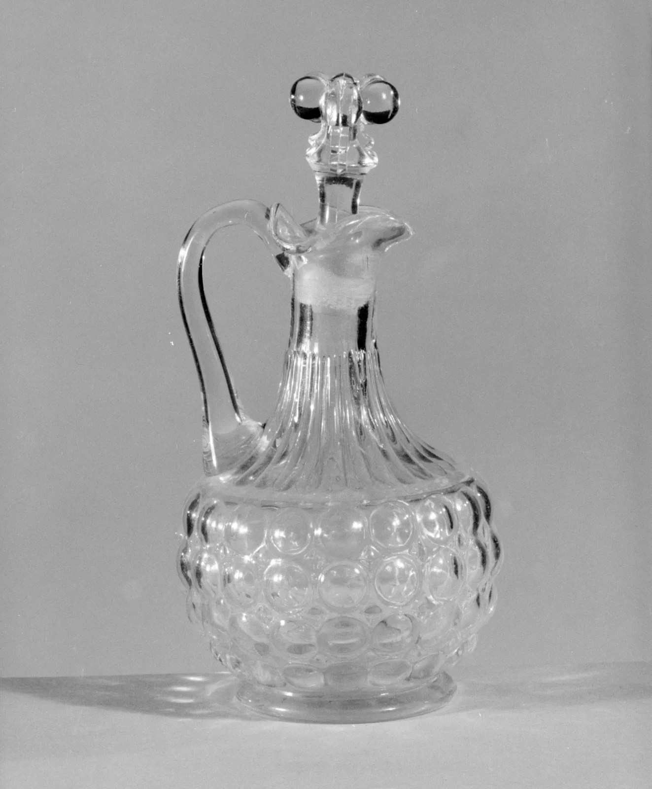 Clear glass cruet bottle with a globular lower body moulded in rows of large round bosses, a plain flared neck, an applied strap handle and a faceted trefoil stopper