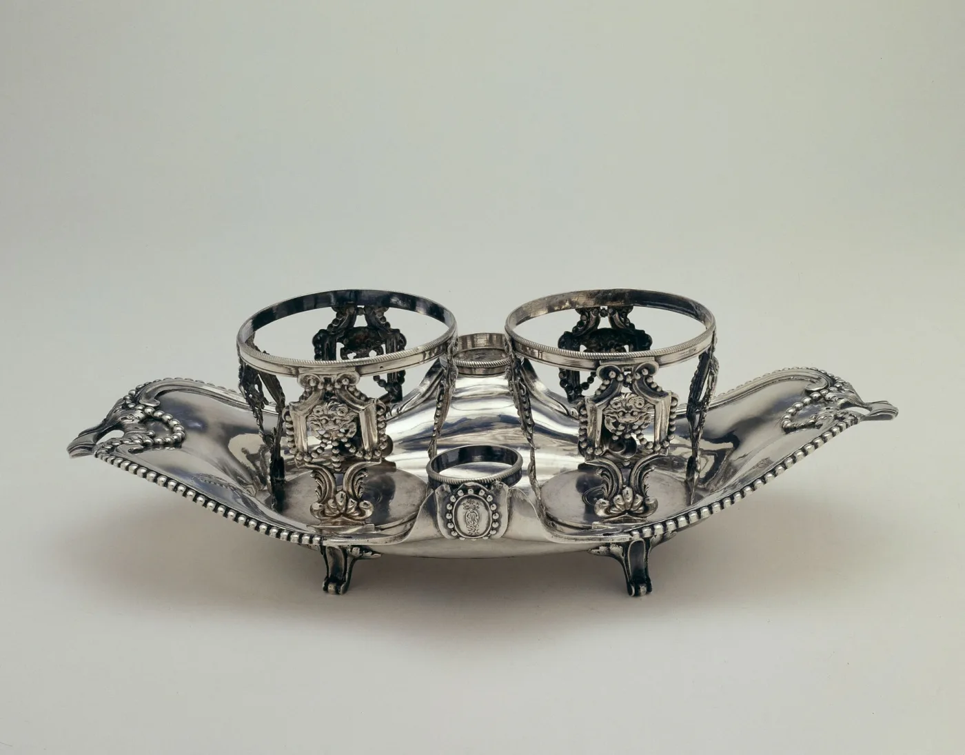 Shallow boat-shaped silver stand with a beaded wavy rim, two tall pierced ring holders with applied medallions flanking a smaller low ring at the centre, standing on four cast feet