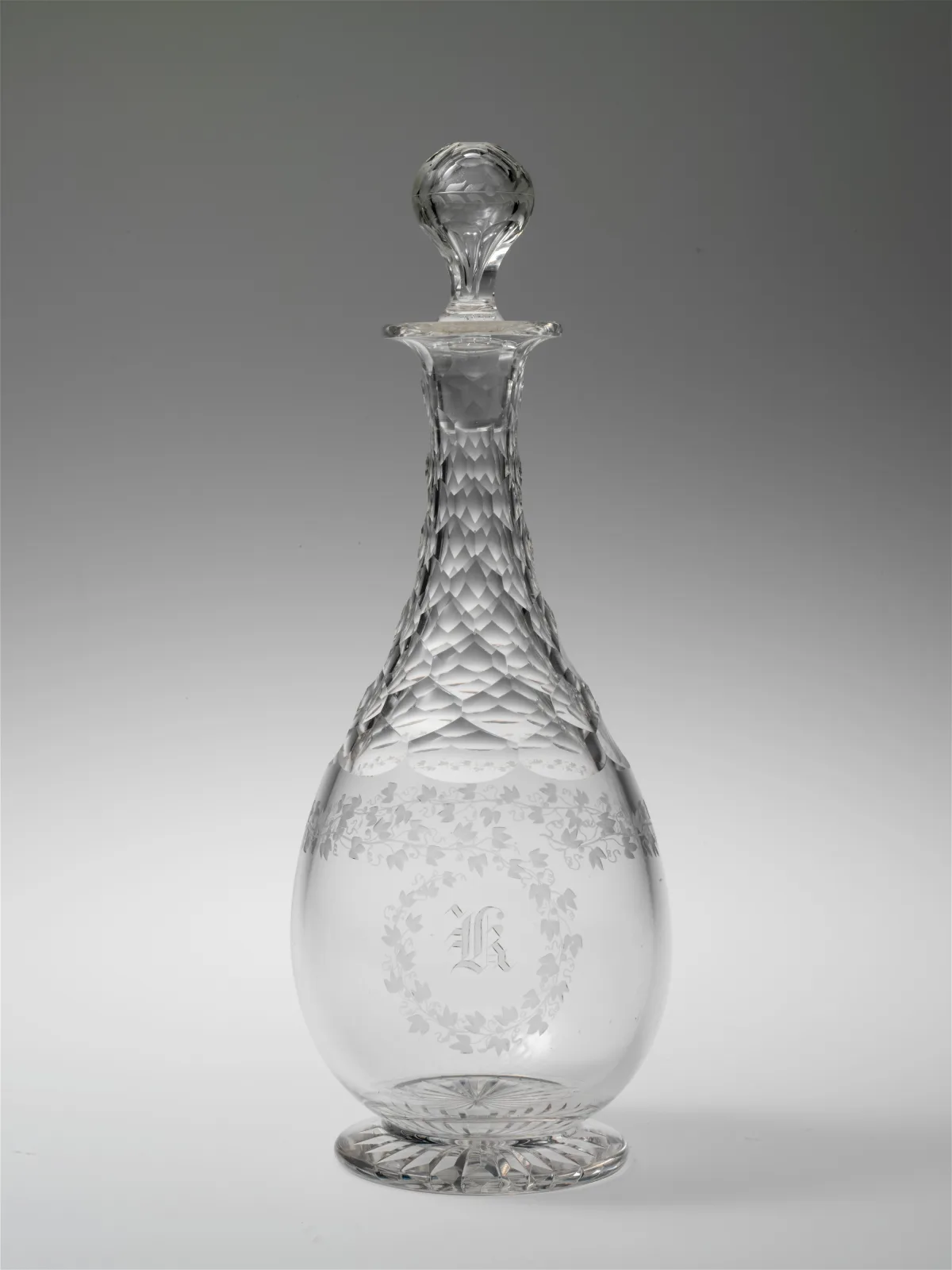 Tall pear-shaped decanter with a long neck cut in overlapping scales, an engraved band of ivy round the body enclosing a monogram in a wreath, a cut star under the foot and a faceted ball stopper on a slim frosted plug