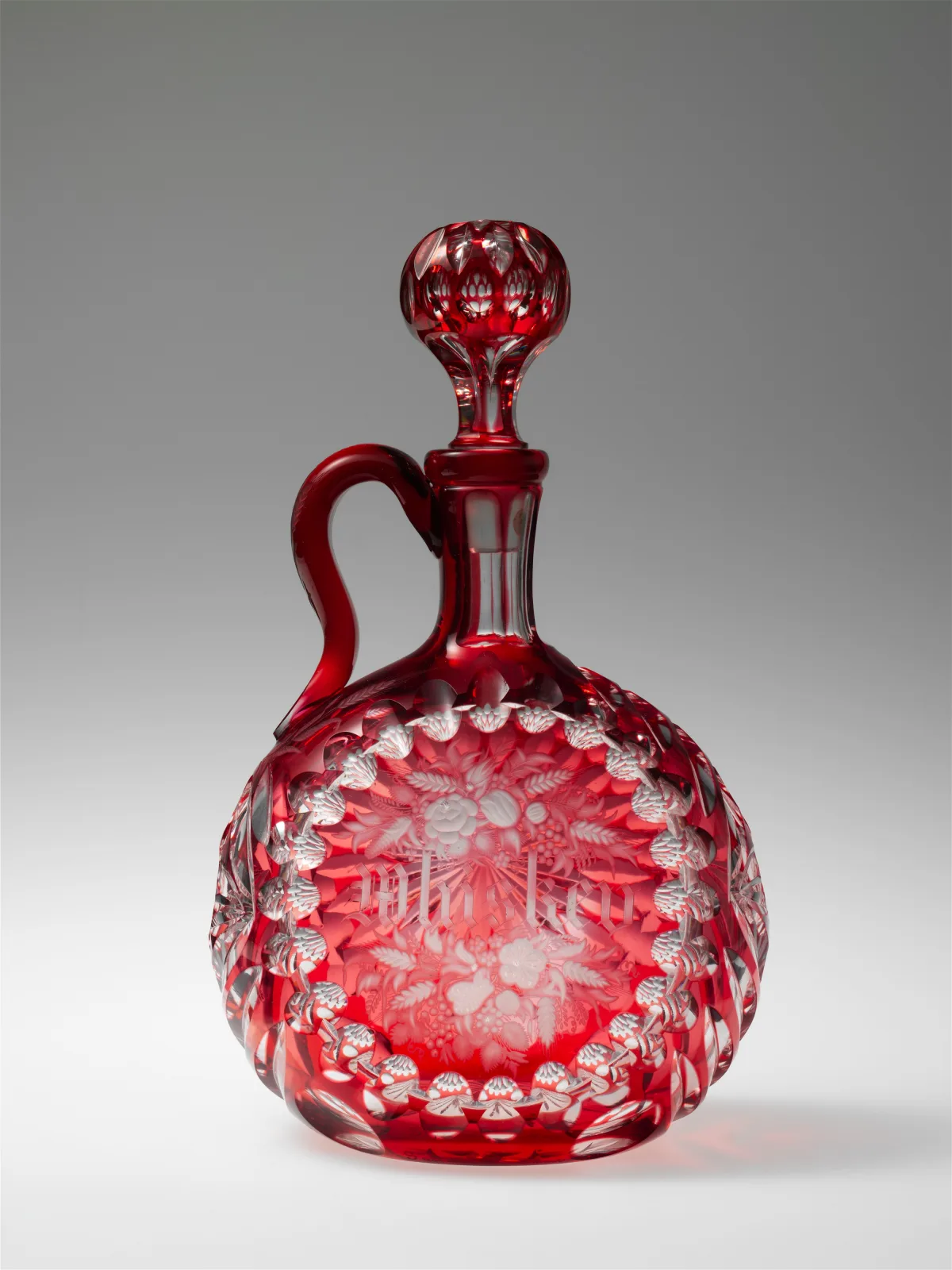 Round-bodied ruby glass decanter with an applied ruby handle, its lower body cut through the red layer to clear glass in a band of pointed ovals and a flower spray, with a cut ruby ball stopper