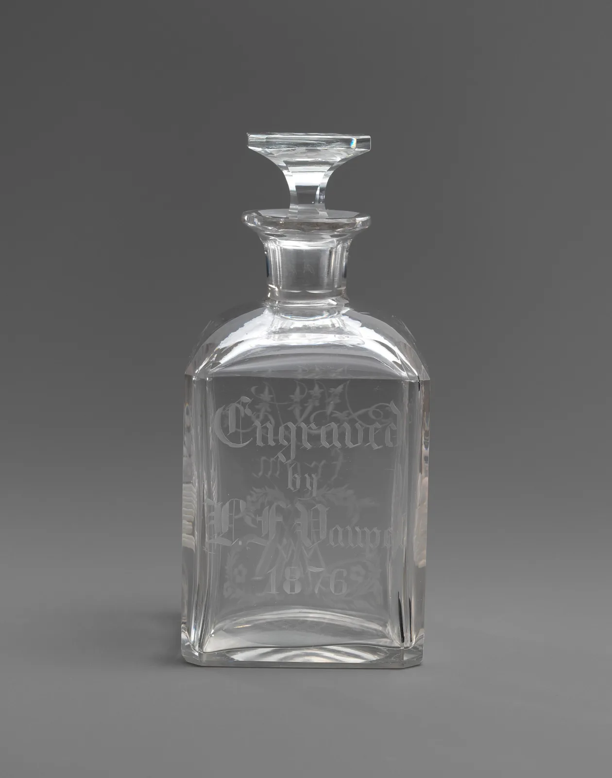 Square-sectioned clear glass bottle with sloping shoulders and a flat rectangular stopper, its front panel engraved in blackletter script reading Engraved by, a personal name, and the date 1876, surrounded by fine engraved foliage