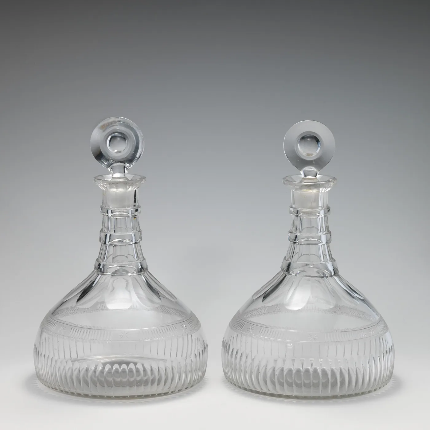 Two matching broad-based decanters standing side by side, each with a low conical body cut with vertical flutes, three rings on a short neck and a flat disc stopper
