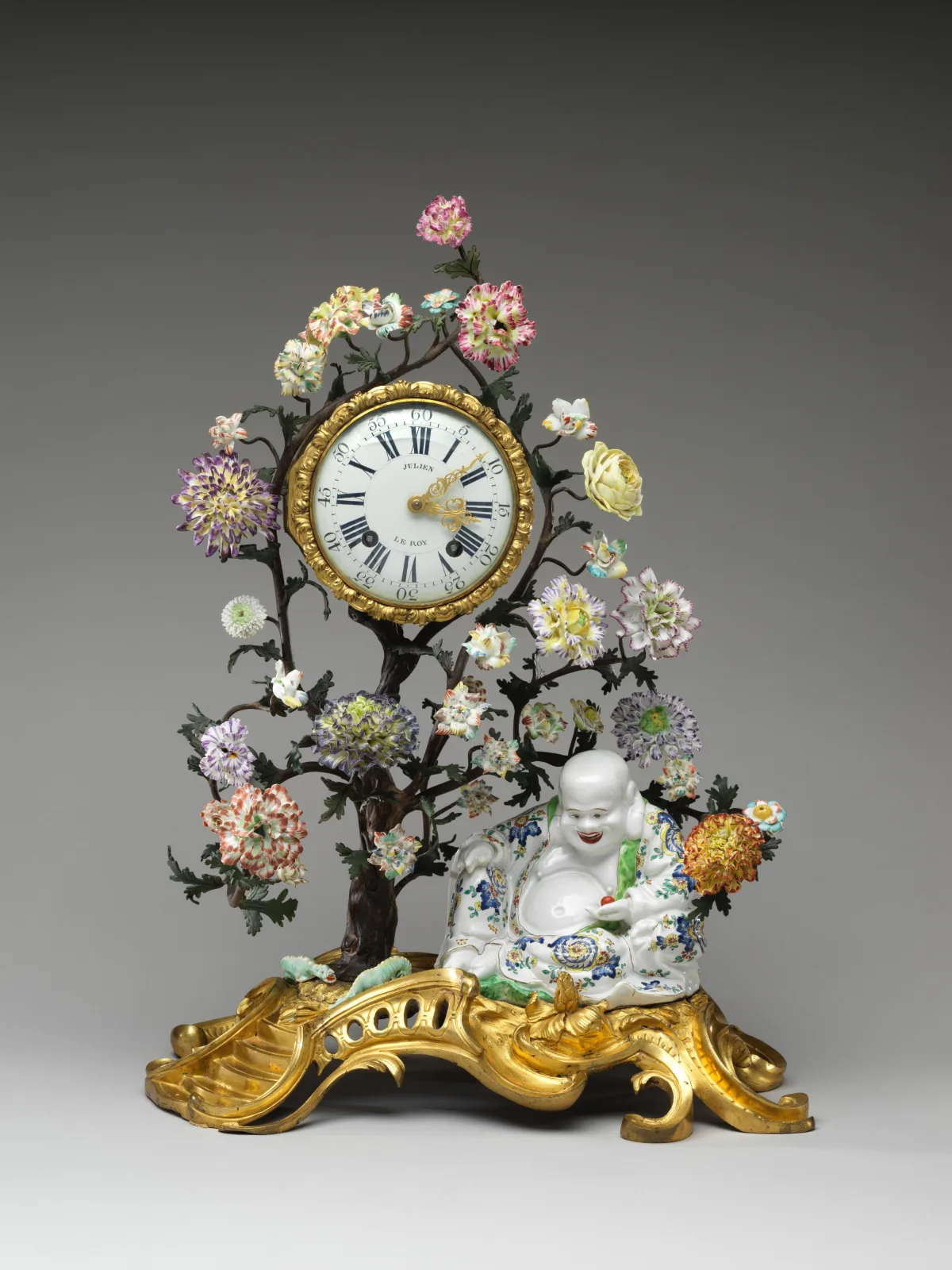 Clock whose enamel dial is set into the trunk of a porcelain tree covered in coloured flowers, with a seated laughing figure in enamelled porcelain below, the whole standing on an elaborate gilt-bronze rococo base